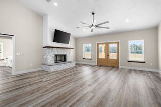 Spacious living room with fireplace, TV, wood floors, and light-colored walls. Double doors and windows provide natural light.