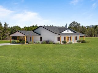 White house with black roof and carport, set on a grassy lawn under a blue sky.