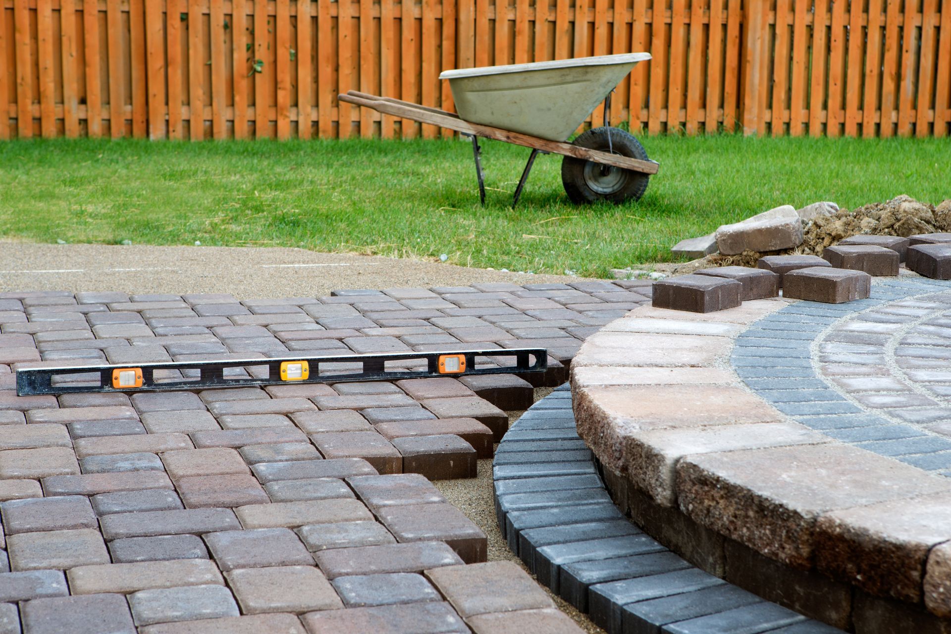 A partially constructed brick patio with a level, wheelbarrow, and wooden fence in the background.
