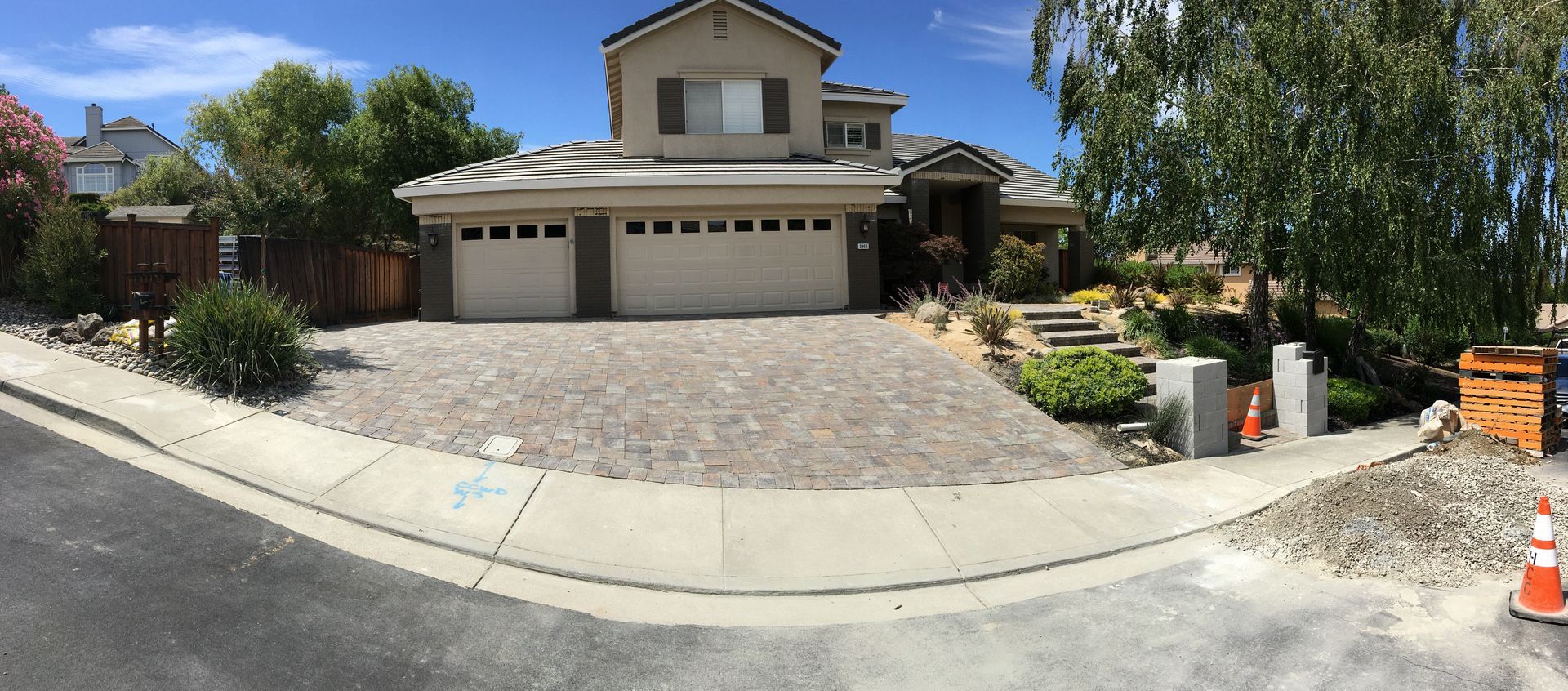 A house with a cobblestone driveway, surrounded by a sidewalk and trees. Construction materials are visible.