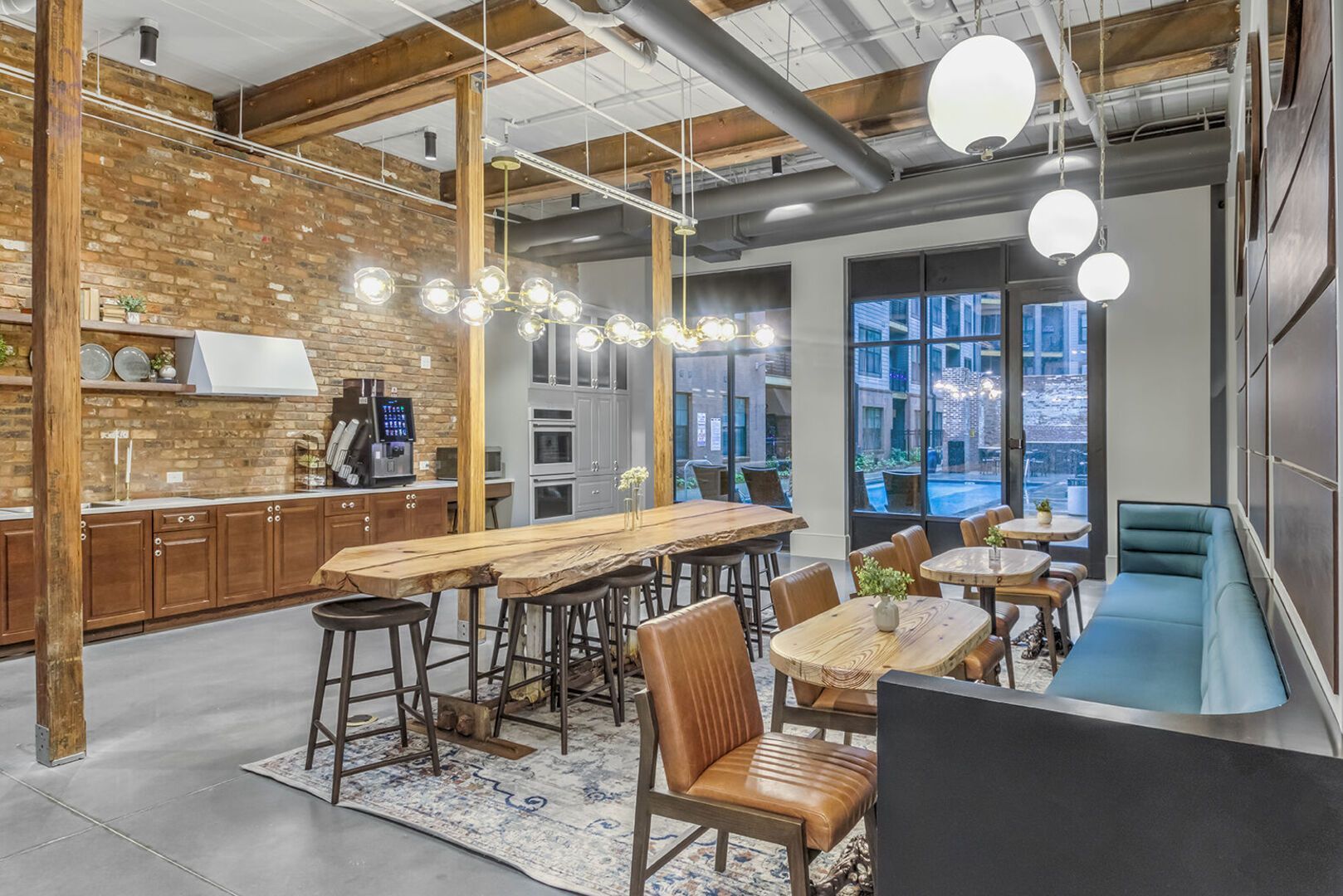 Industrial-style communal kitchen and lounge with a long wooden table, stools, brick wall, and large windows.