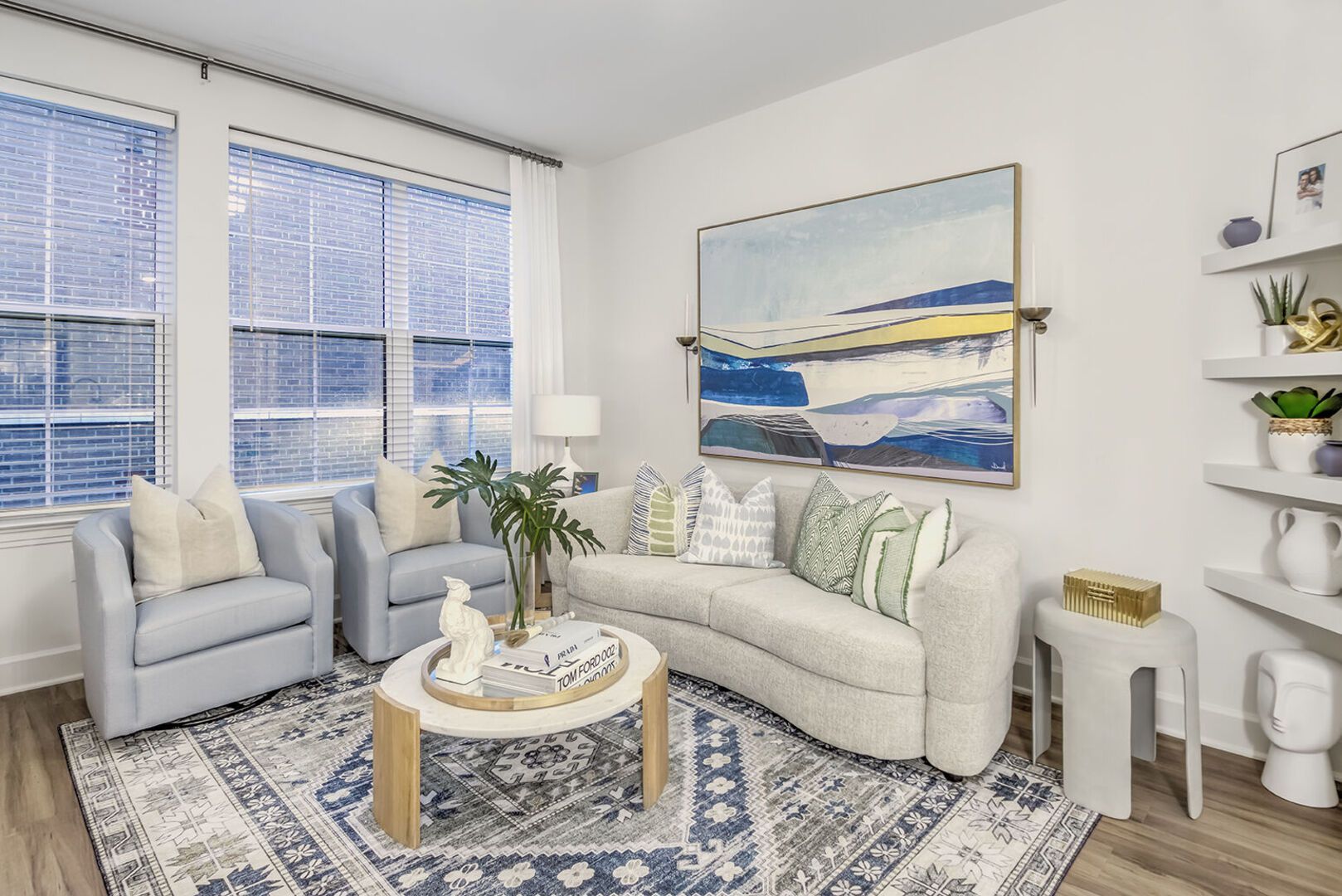 Bright living room in a modern apartment with a light sofa, two blue chairs, large windows, and abstract wall art.