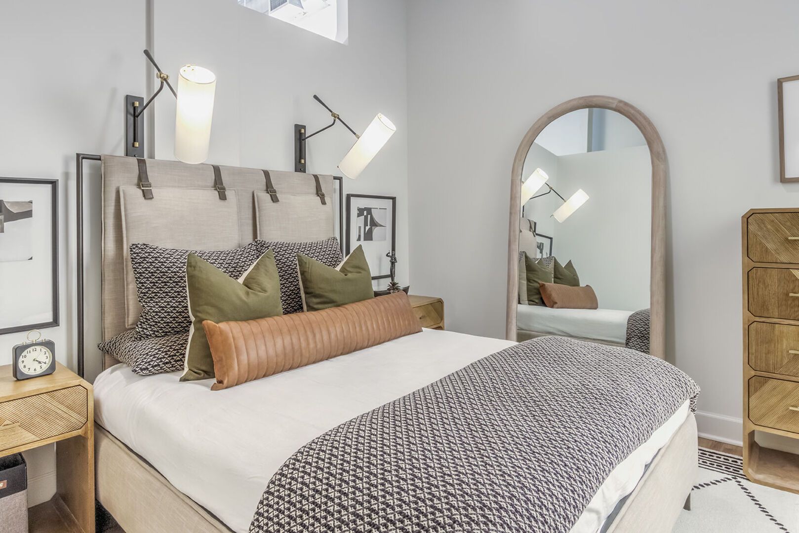 Modern apartment bedroom with a made bed, decorative pillows, and a large arched mirror.