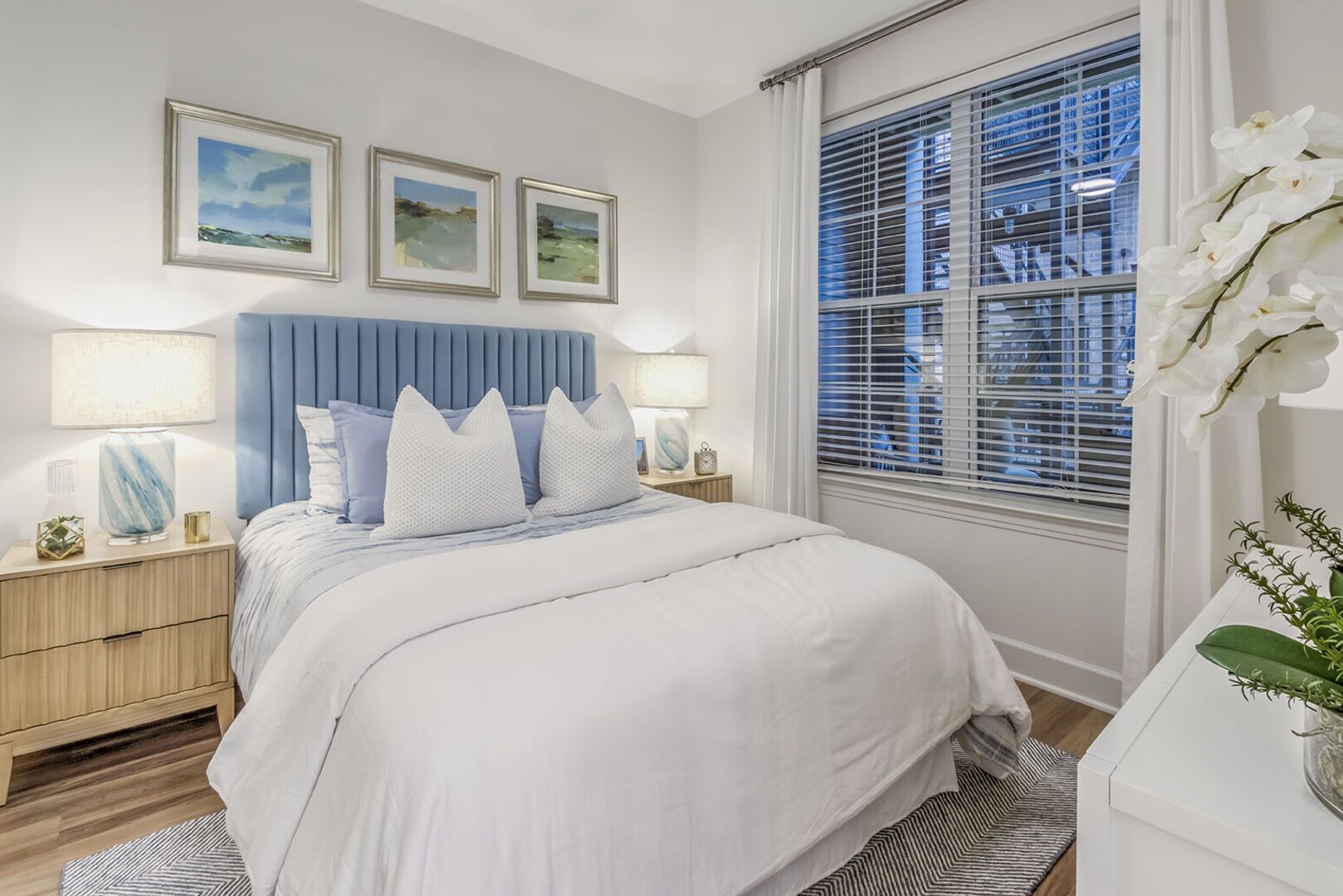 Cozy modern bedroom with a blue upholstered headboard, white bedding, and a large window with blinds.