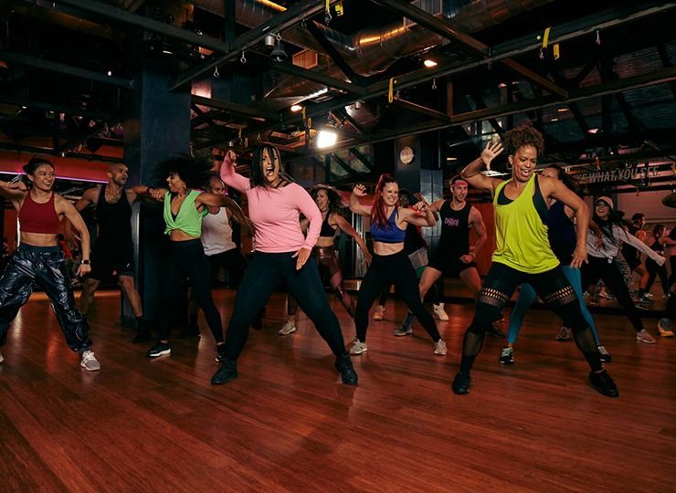 People dancing in a studio, with wooden floor. They wear workout clothes and move with energy.