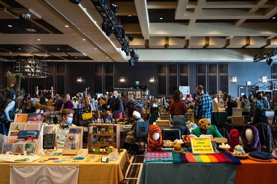 People browsing a vendor's table at an outdoor market, looking at colorful items and jars.
