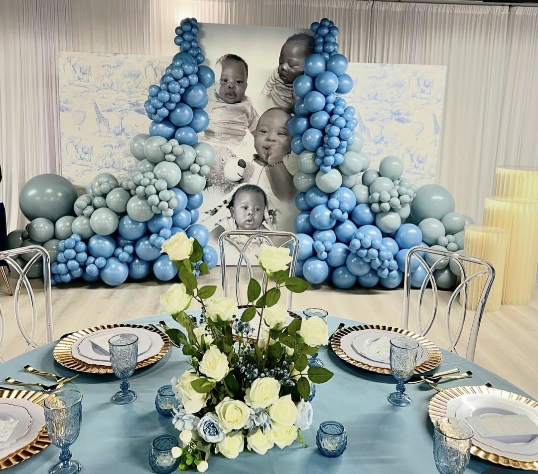 A table with plates , glasses , and flowers in front of a wall of blue balloons.