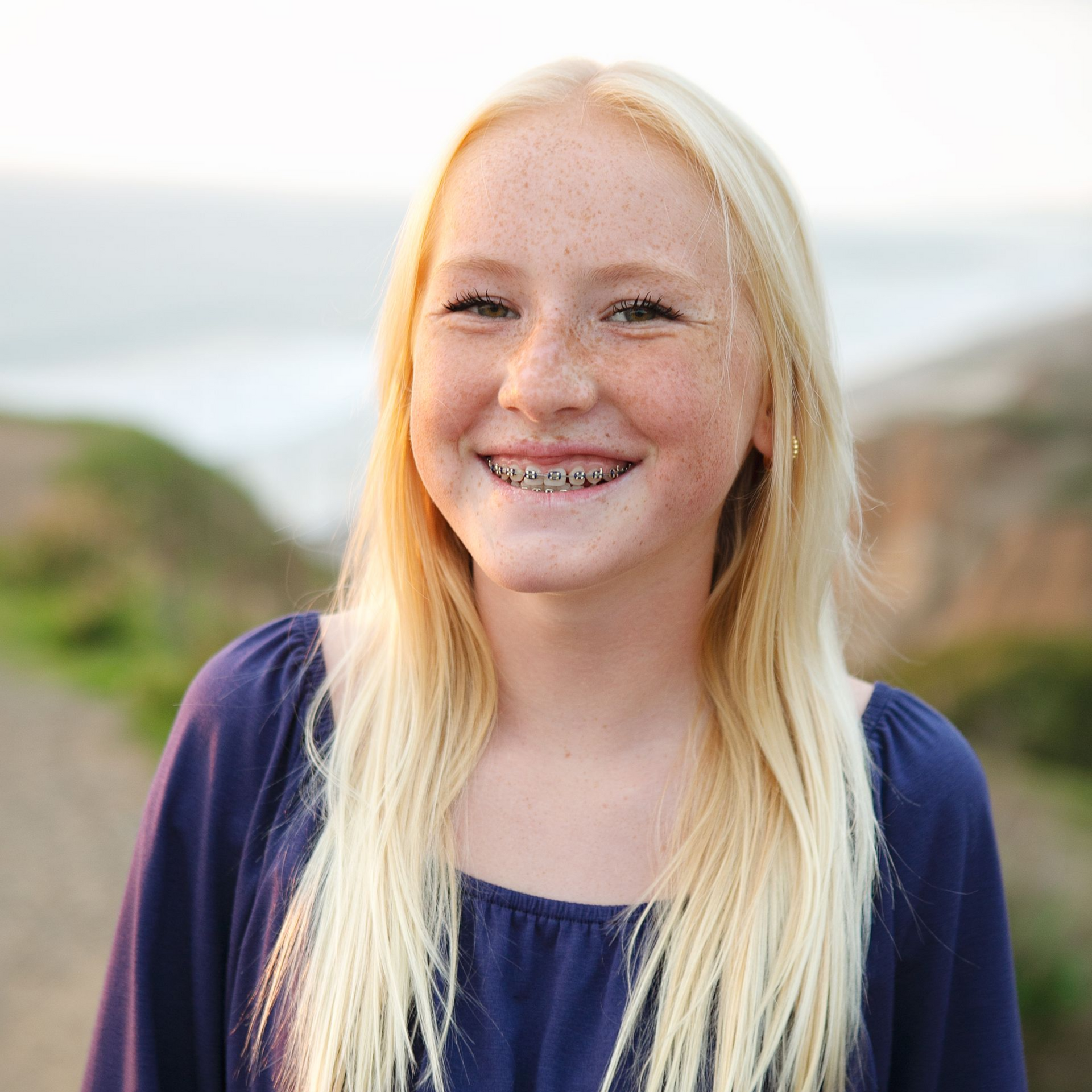 Blonde person with braces smiles at the camera, wearing a blue top, with a blurred outdoor background.
