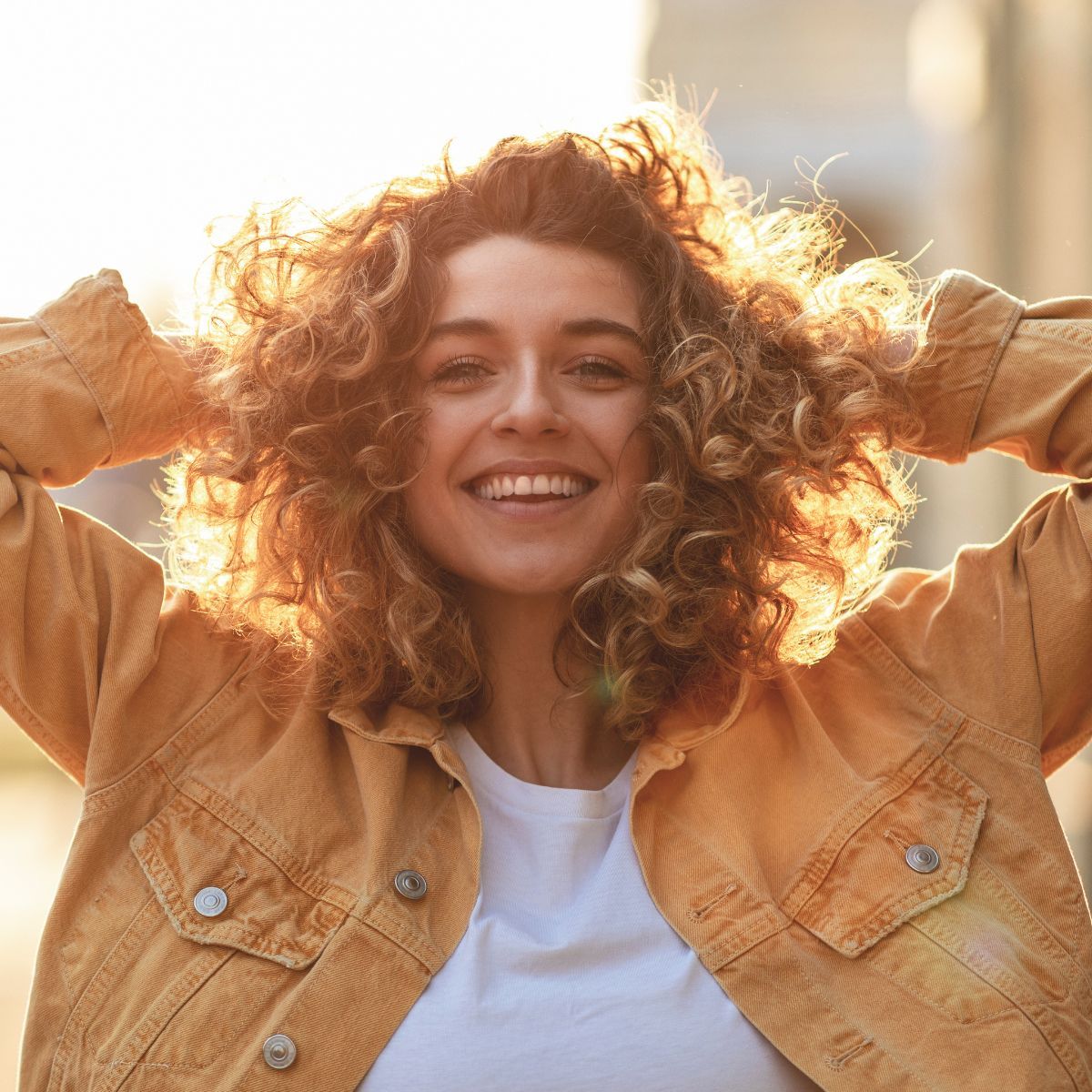 A woman with curly hair is wearing a yellow jacket and smiling.