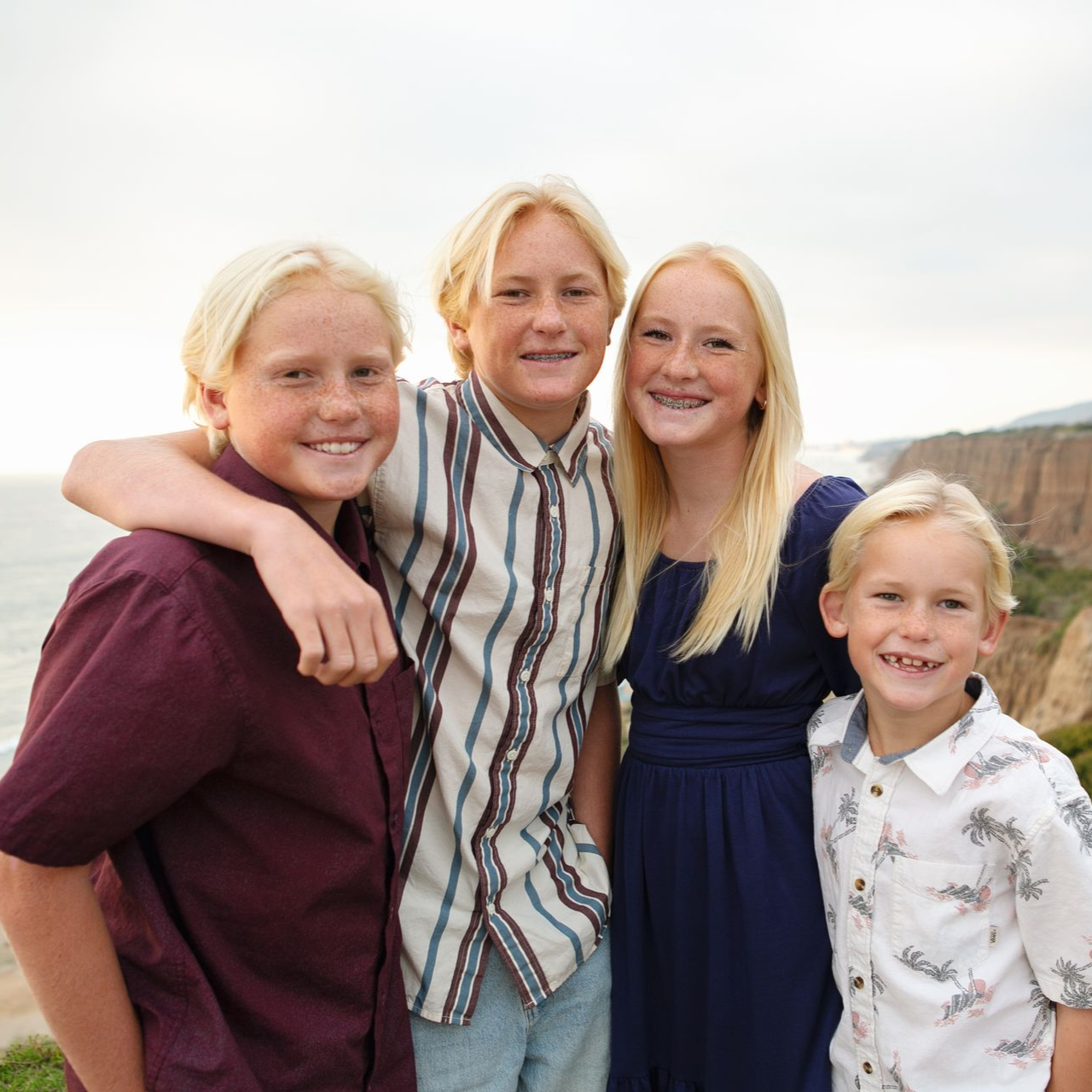 Four blonde children smiling, posing together outdoors by the ocean.
