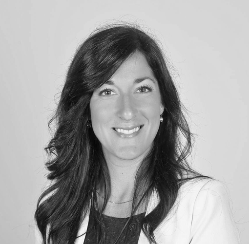 Femme souriante aux cheveux foncés portant un blazer blanc et un haut foncé, sur un fond clair.