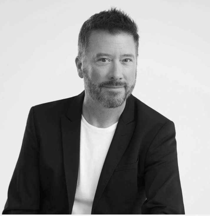 Un homme en veste de costume noire et chemise blanche, regardant la caméra avec un léger sourire.