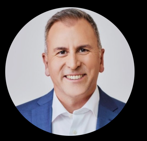 Une femme aux cheveux bouclés sourit, portant un blazer clair sur un haut foncé, sur fond gris.