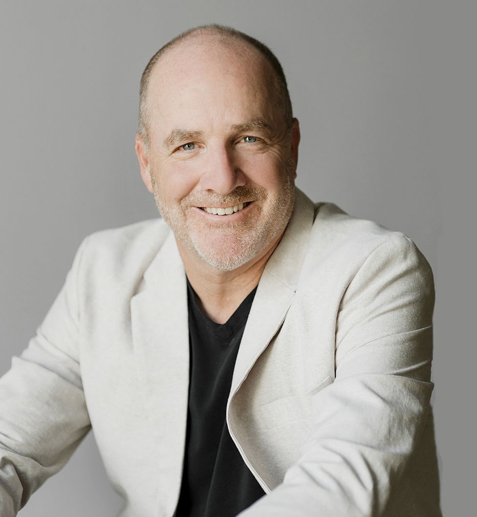 Homme souriant, vêtu d'un blazer léger et d'une chemise noire, sur fond gris.