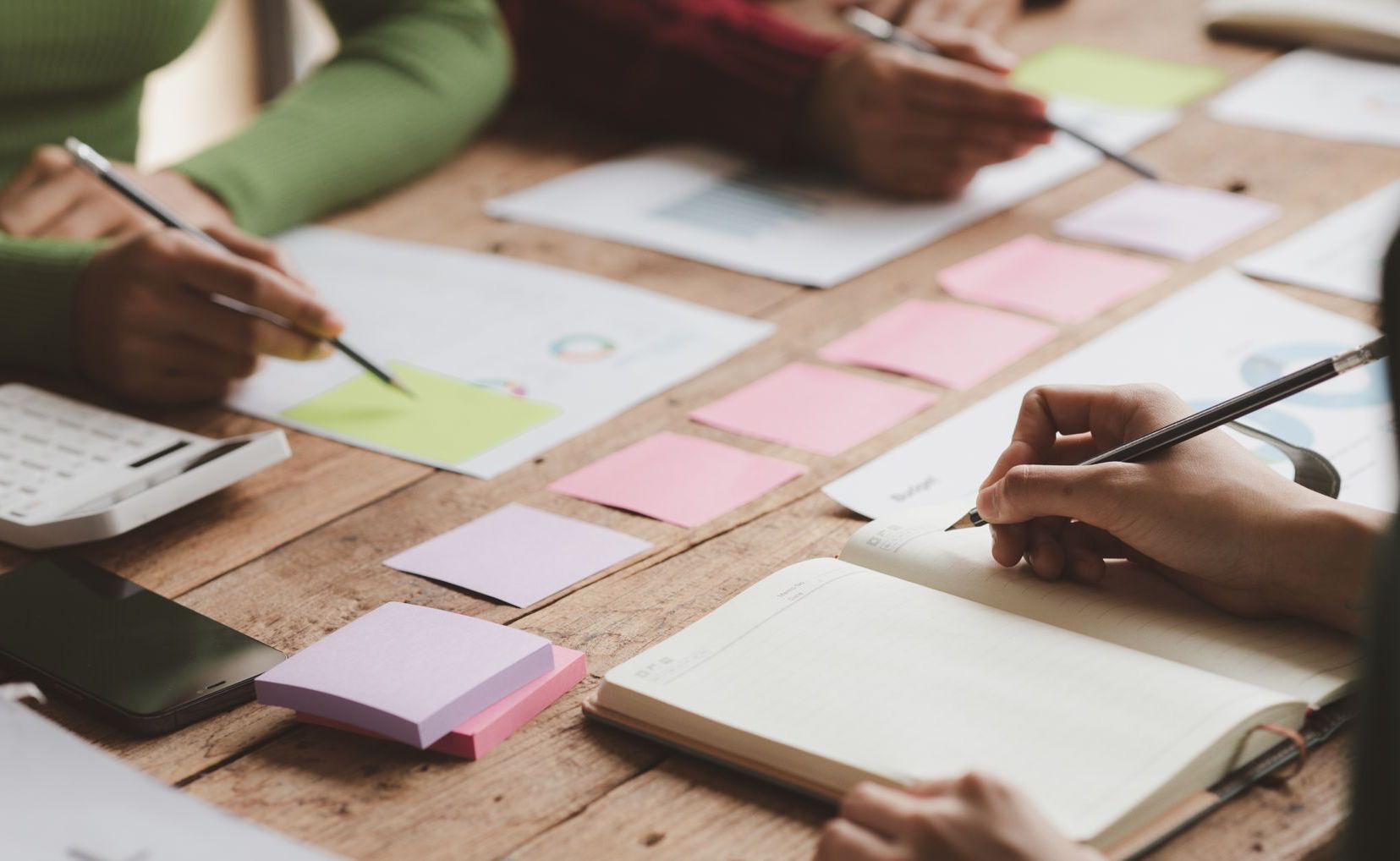 Des gens écrivent et travaillent à une table en bois, avec des papiers, des notes autocollantes et une calculatrice.