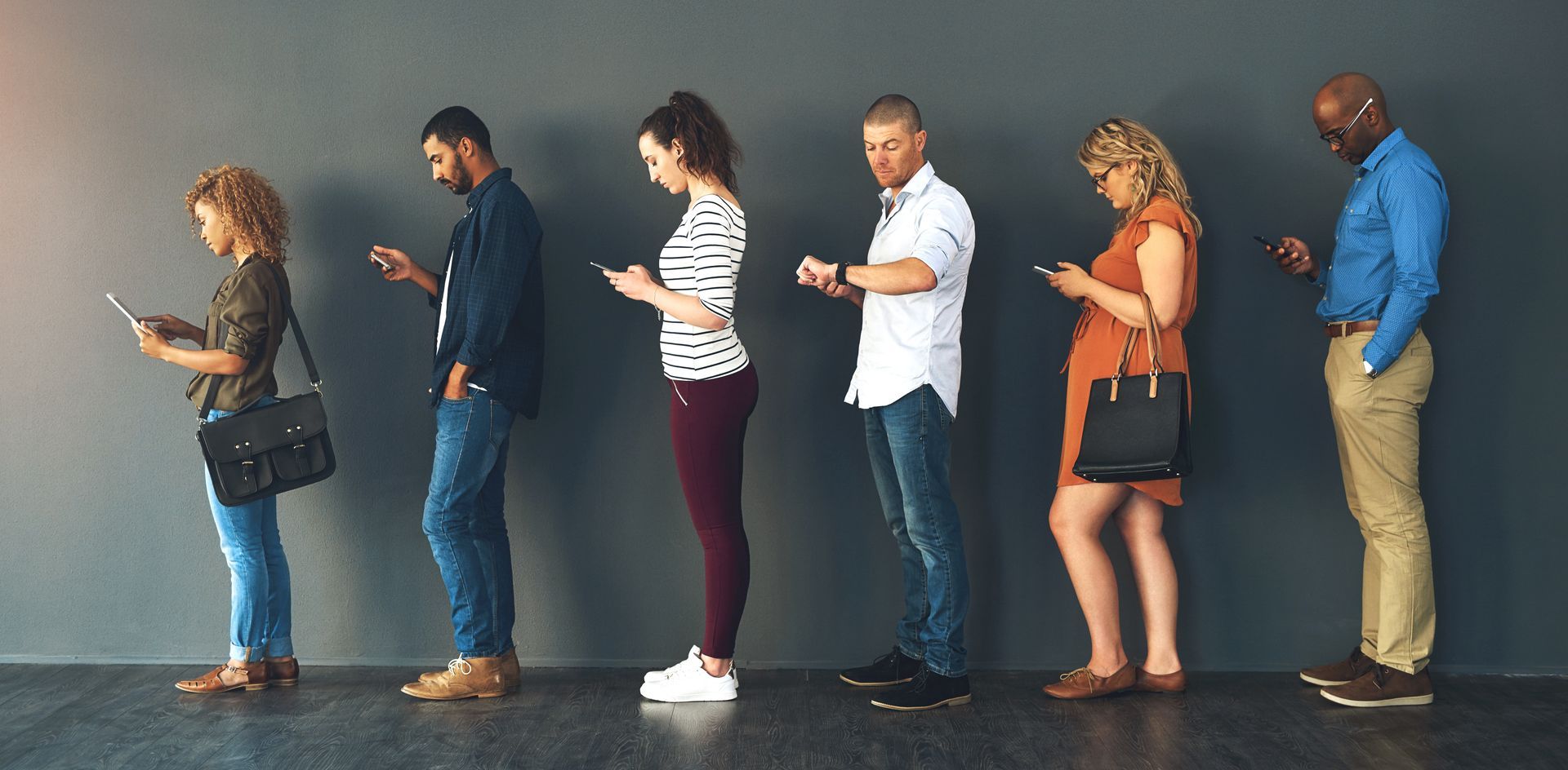 Des gens debout dans une file, chacun regardant un téléphone, contre un mur gris.