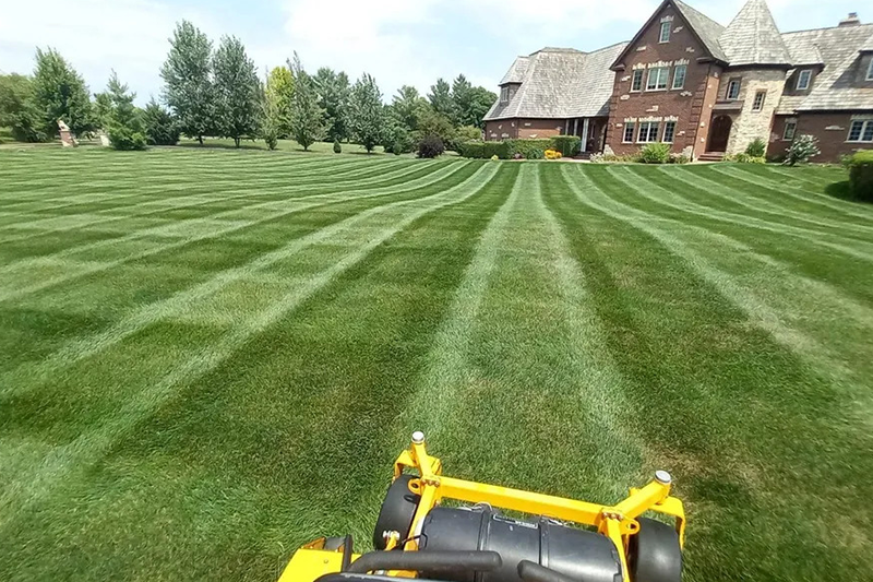A yellow lawn mower is cutting a lush green lawn in front of a large house.