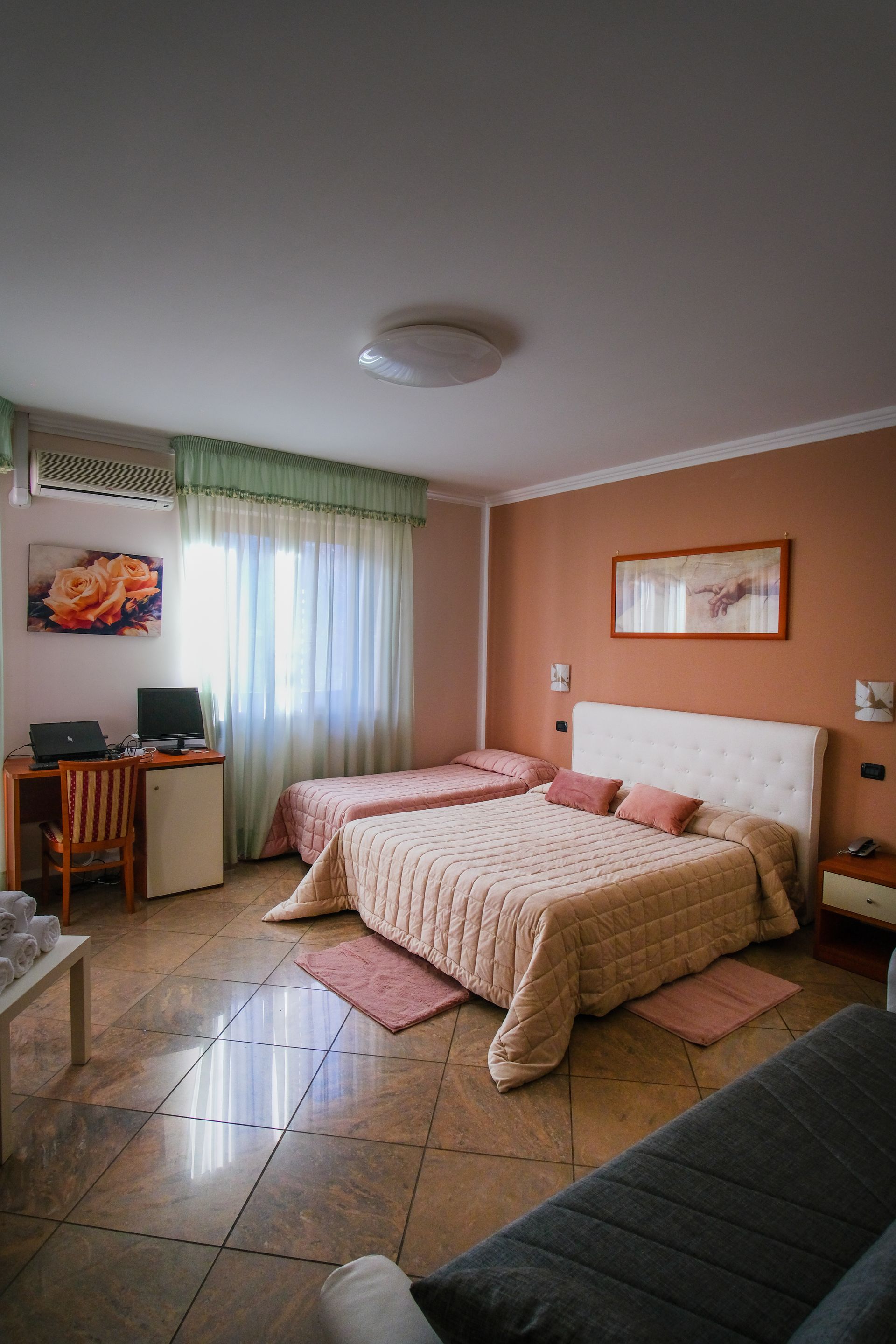 Camera da letto con letto matrimoniale, letto singolo, scrivania e piccolo frigorifero. Tonalità calde e finestra luminosa.