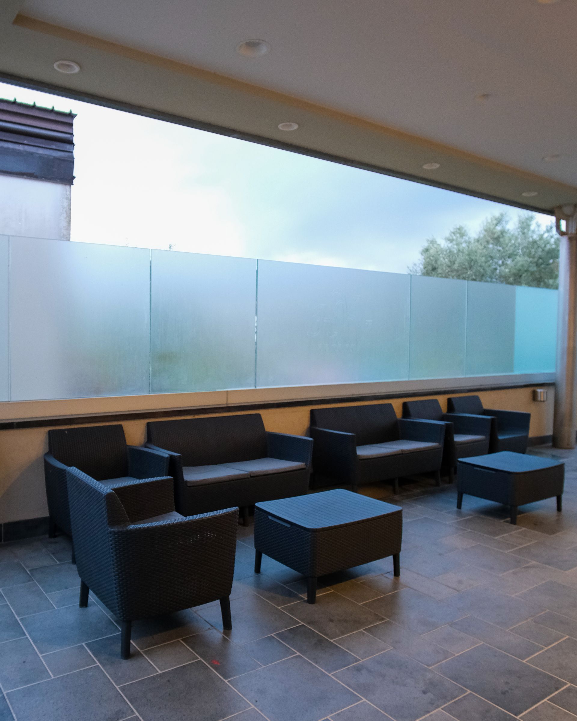 Area salotto all'aperto con mobili scuri, parete in vetro smerigliato e cielo coperto.