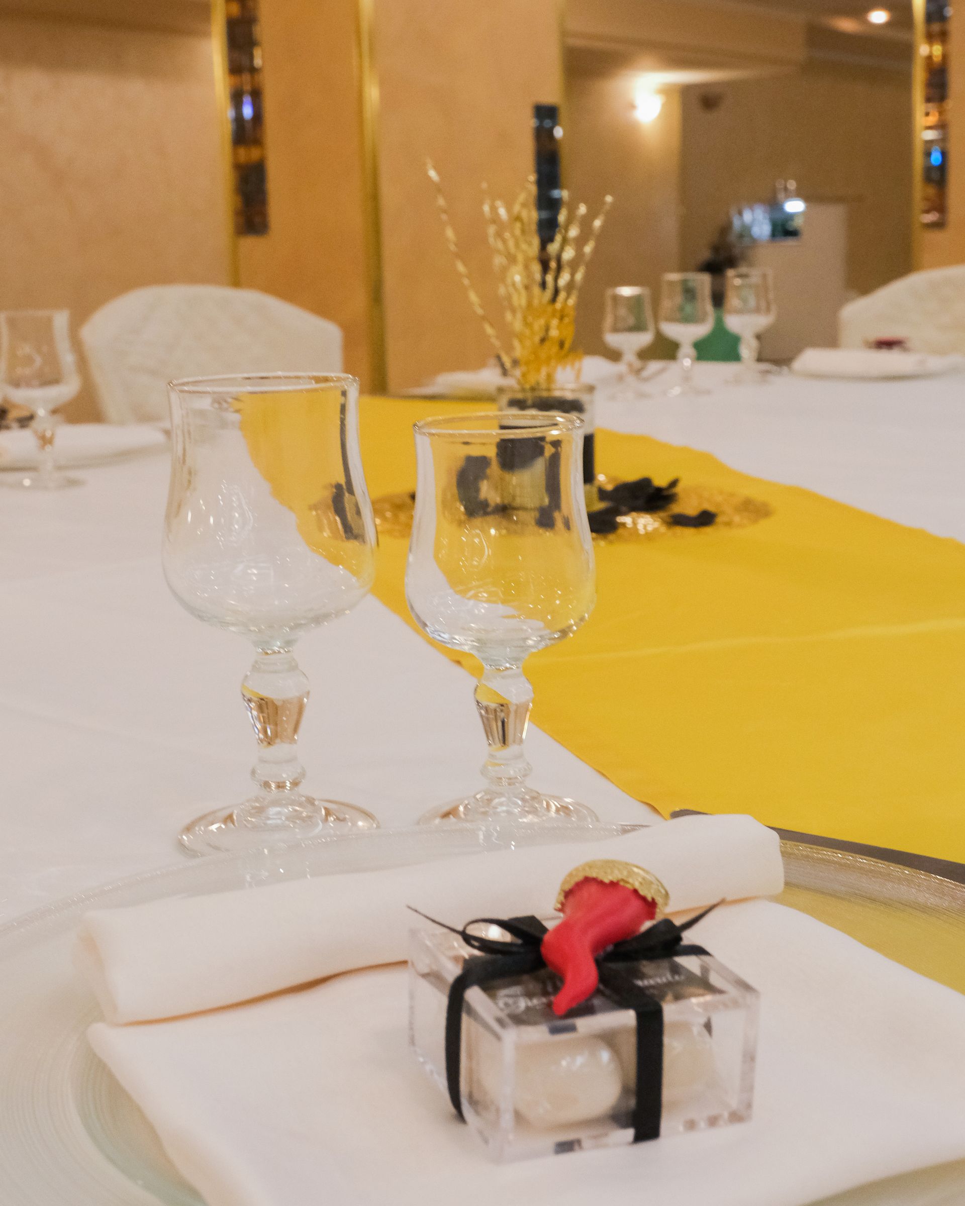 Elegante apparecchiatura con bicchieri da vino, tovagliolo arrotolato e piccola scatola portaconfetti. Runner giallo, tovaglia bianca.