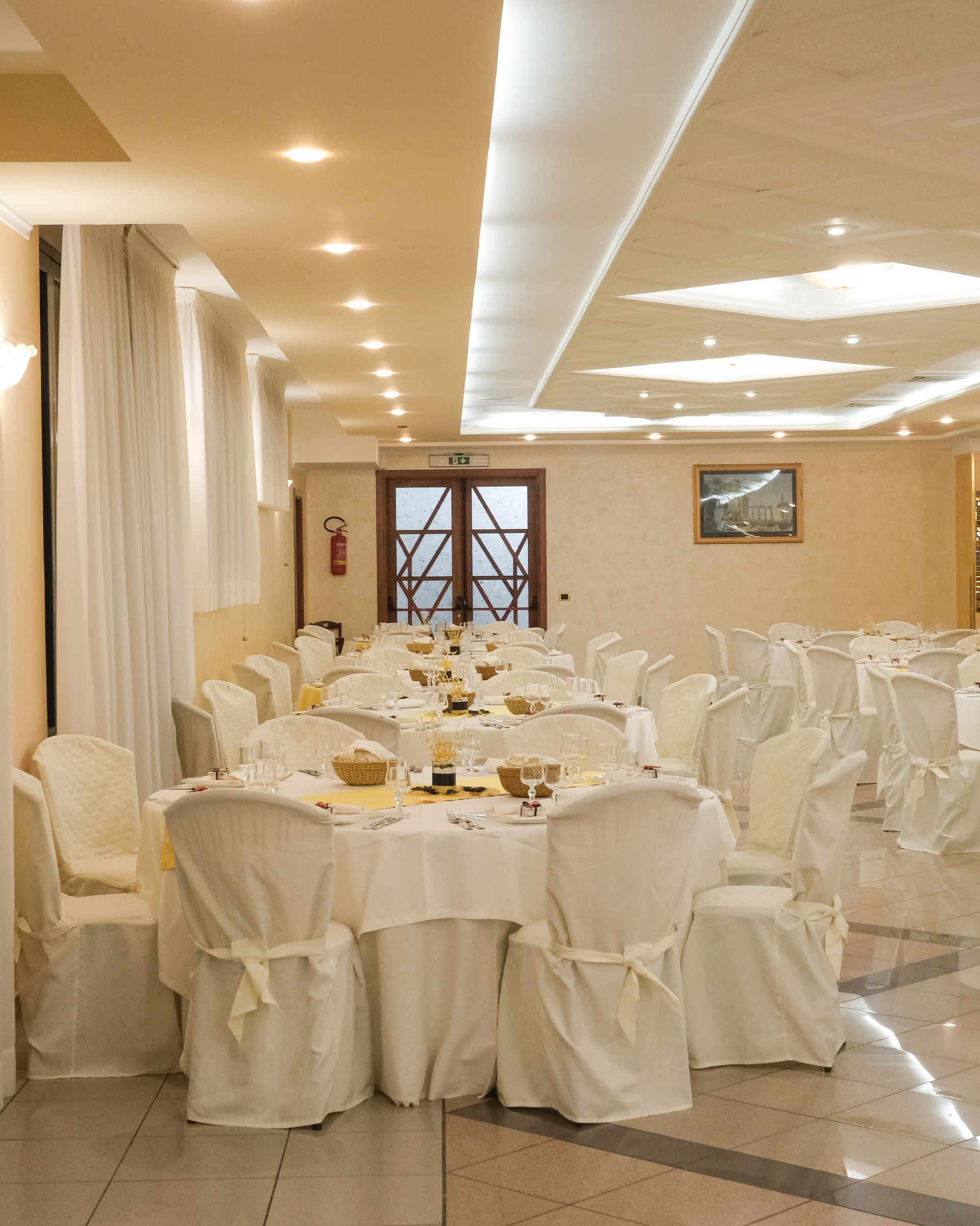 Sala da pranzo con tavoli apparecchiati per un pasto. Tovaglie bianche, sedie e luci soffuse creano un'atmosfera elegante.