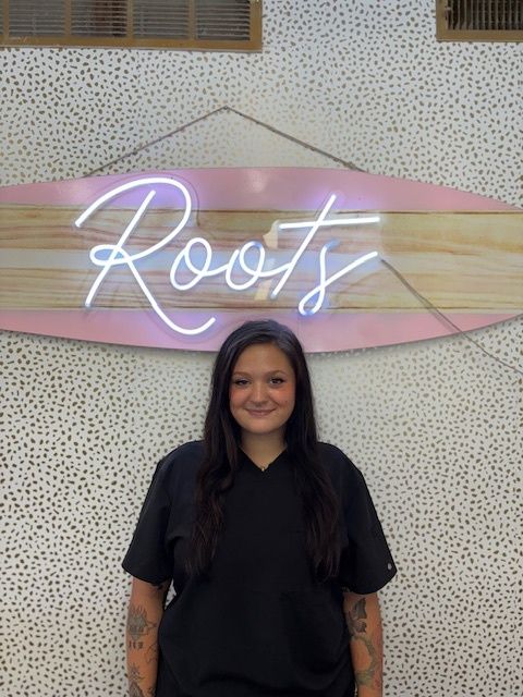 Woman in black scrubs smiles in front of a Roots sign, polka dot wall.