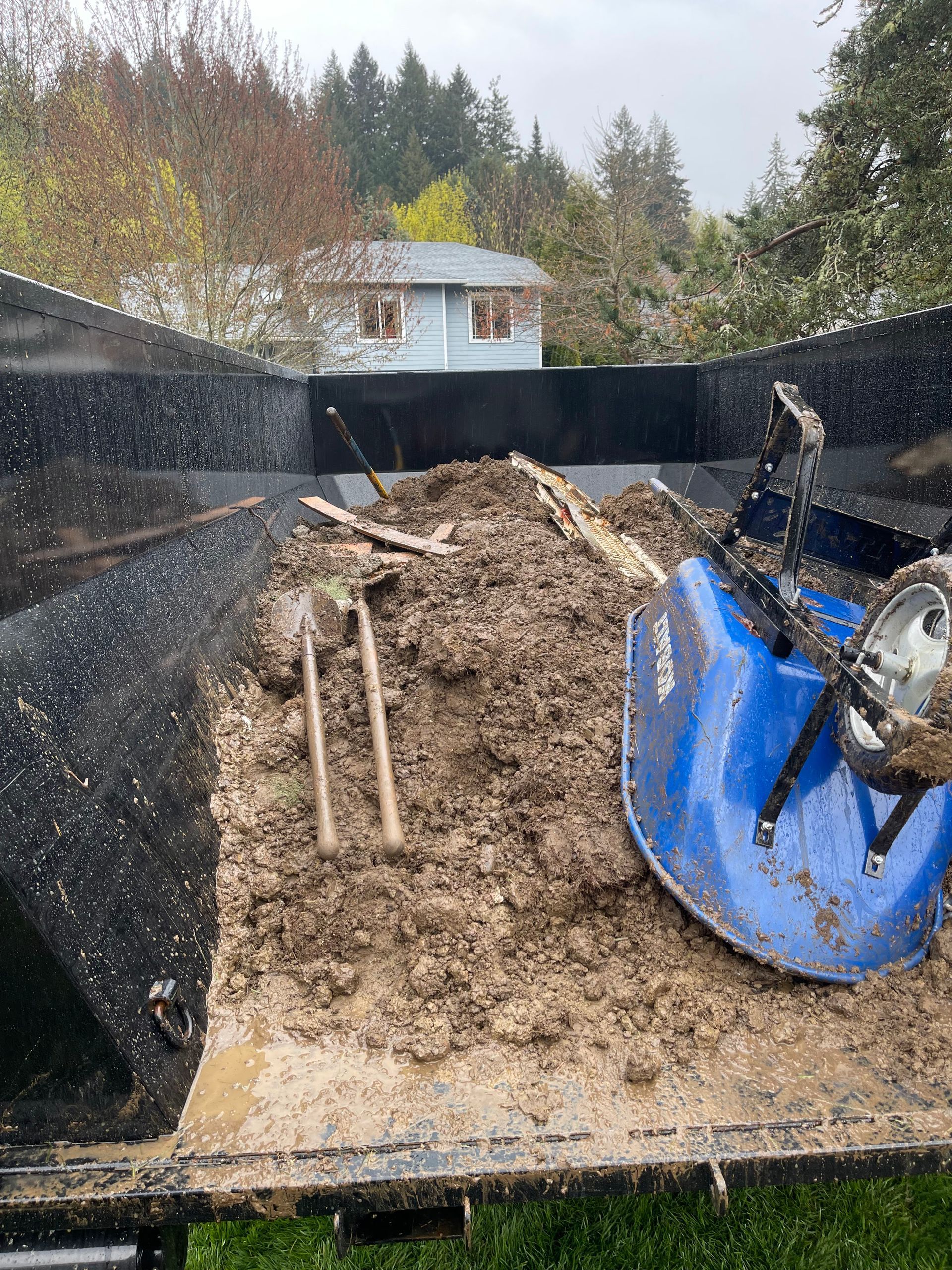 A dump truck bed filled with dirt, tools, and a blue snowblower; outdoor setting.