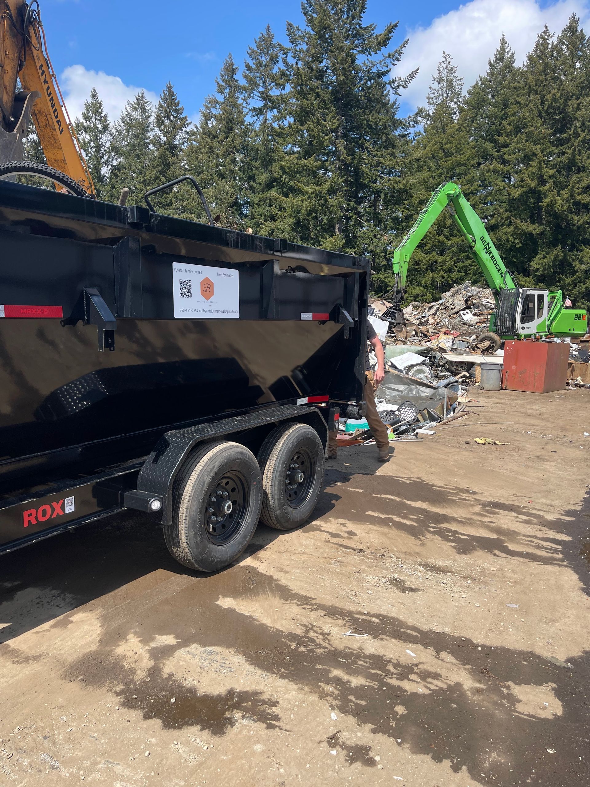 Black trailer in a scrap yard, with a green excavator in the background.