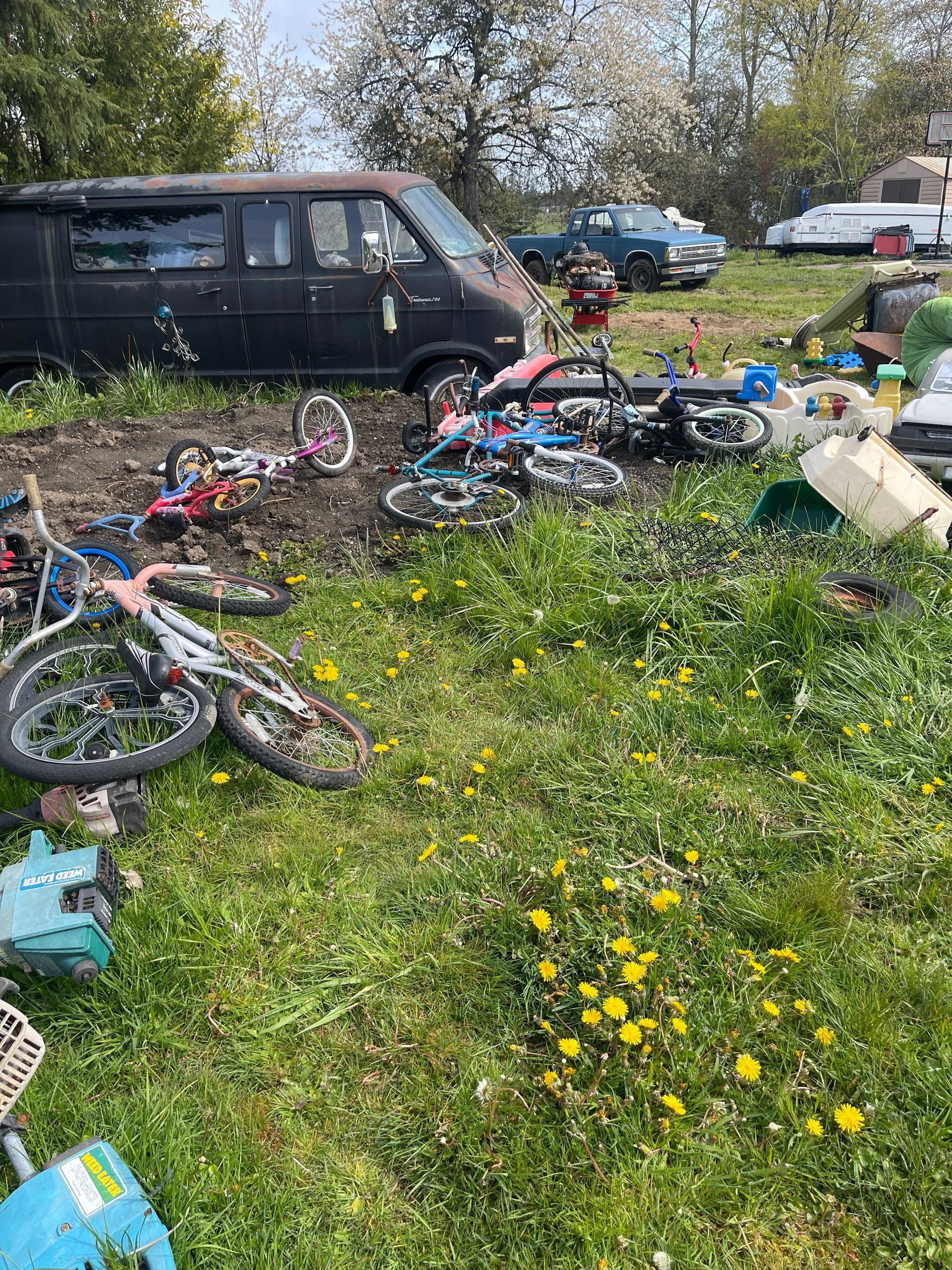 A cluttered yard with a black van, bicycles, and various objects scattered on the grass with yellow flowers.