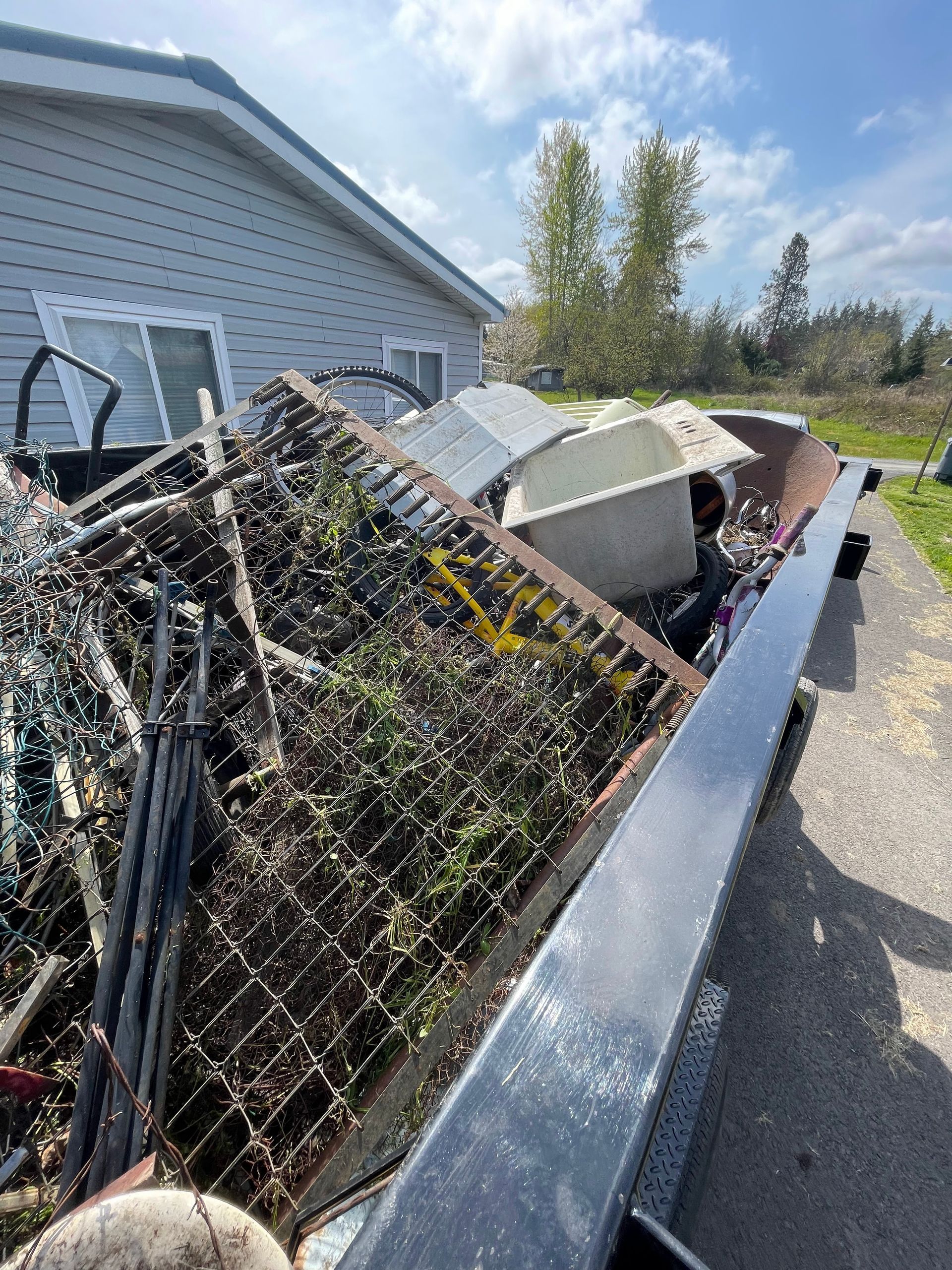 A truck bed filled with scrap metal, a bathtub, and other junk in front of a house on a sunny day.
