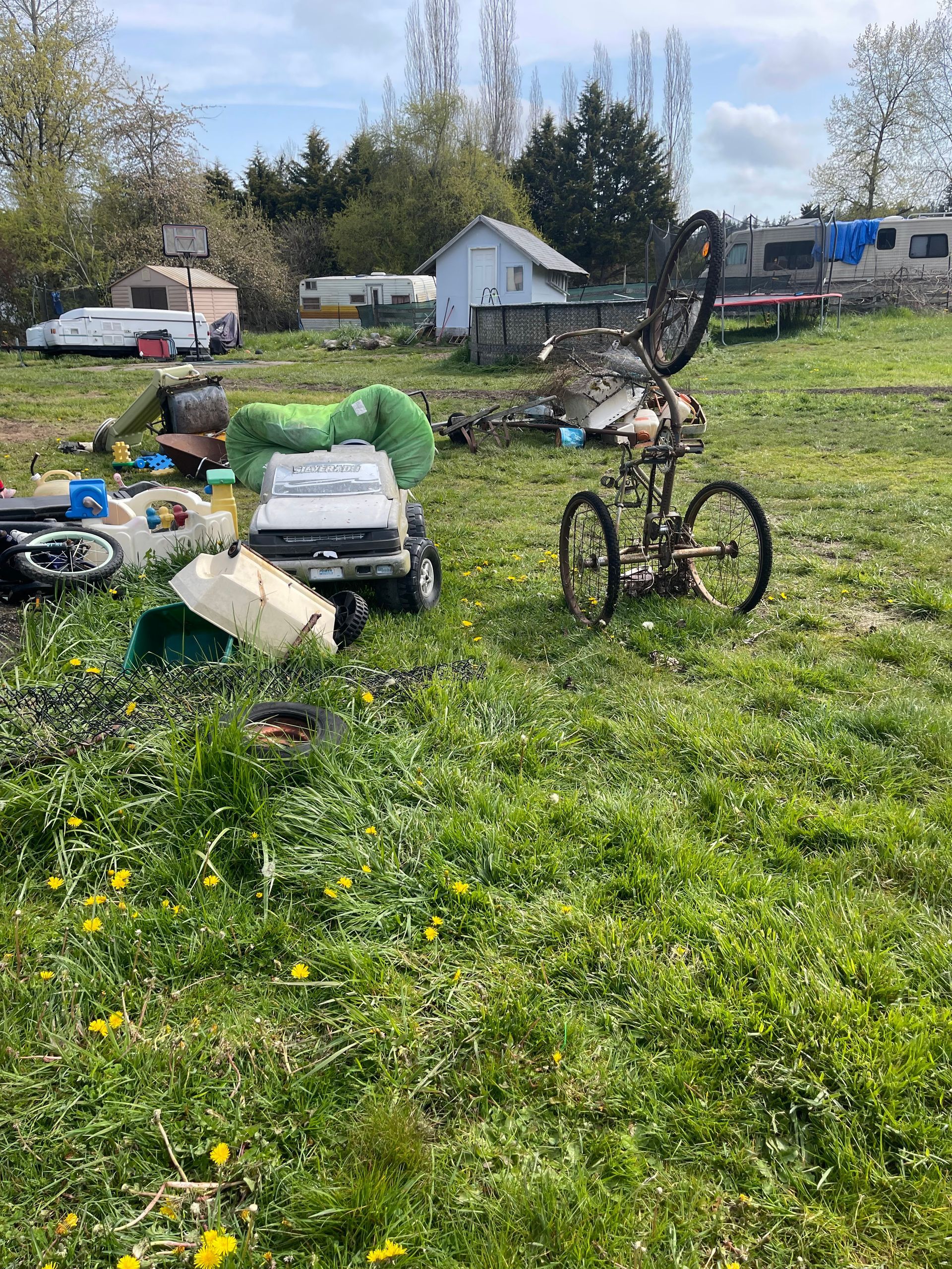 Grassy field with debris including a toy car and old bicycle. Several trailers and a shed are in the background.