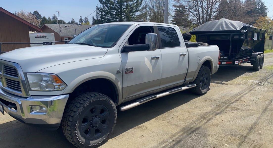 White Ram pickup truck towing a black dump trailer on a gravel road.