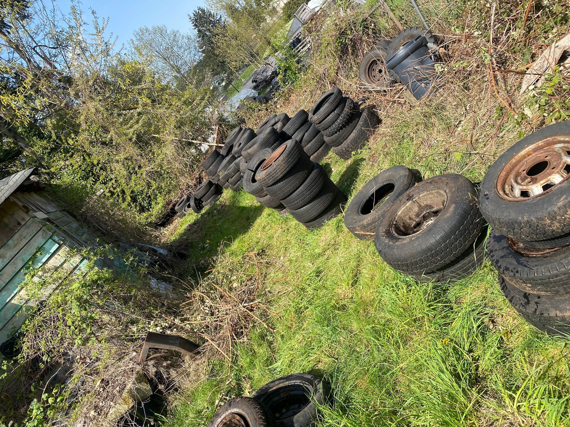 A pile of used tires stacked outdoors on grass. Some tires have rims.