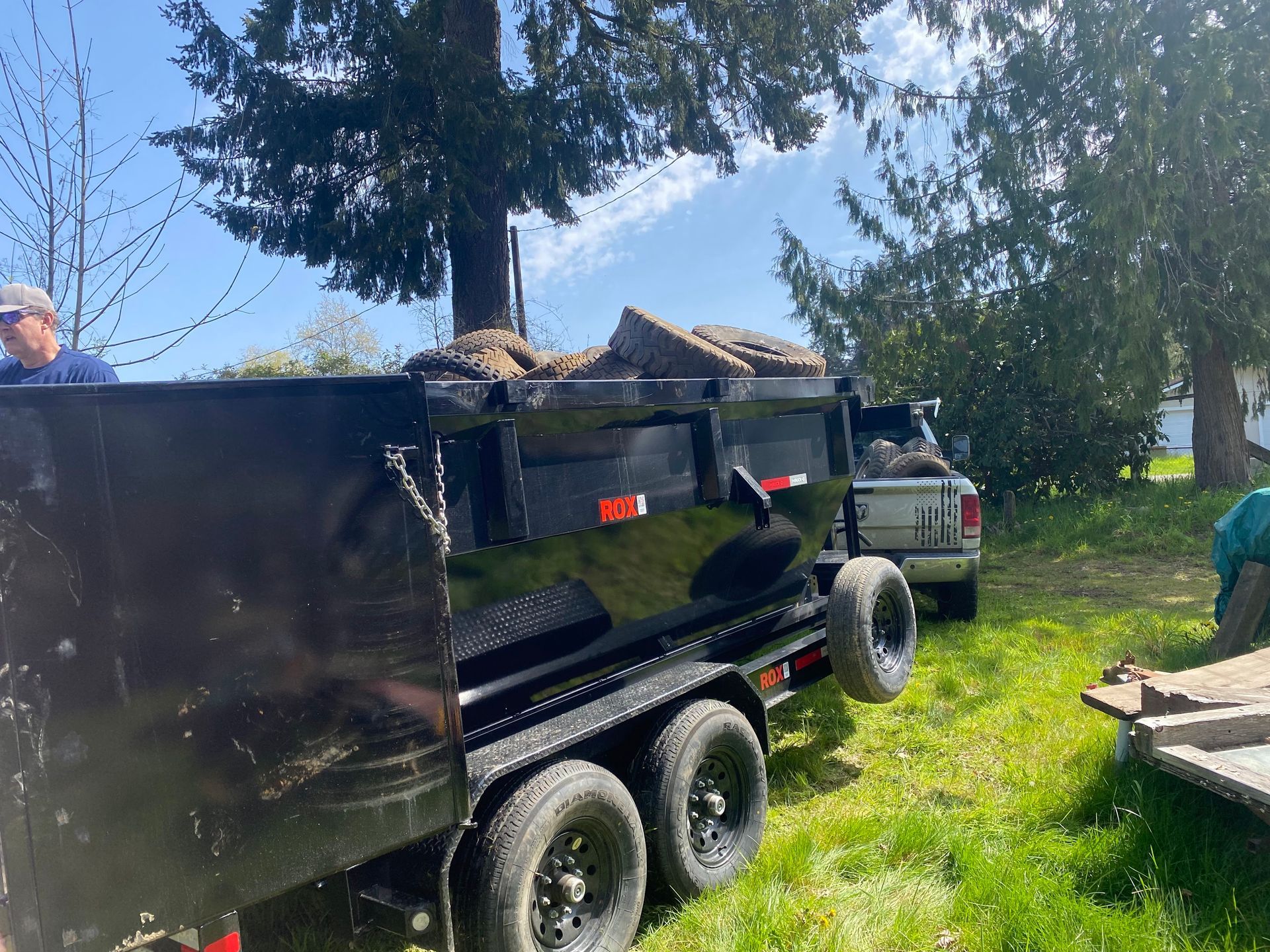 A black dump trailer loaded with debris, next to a truck, on a green lawn with two people.