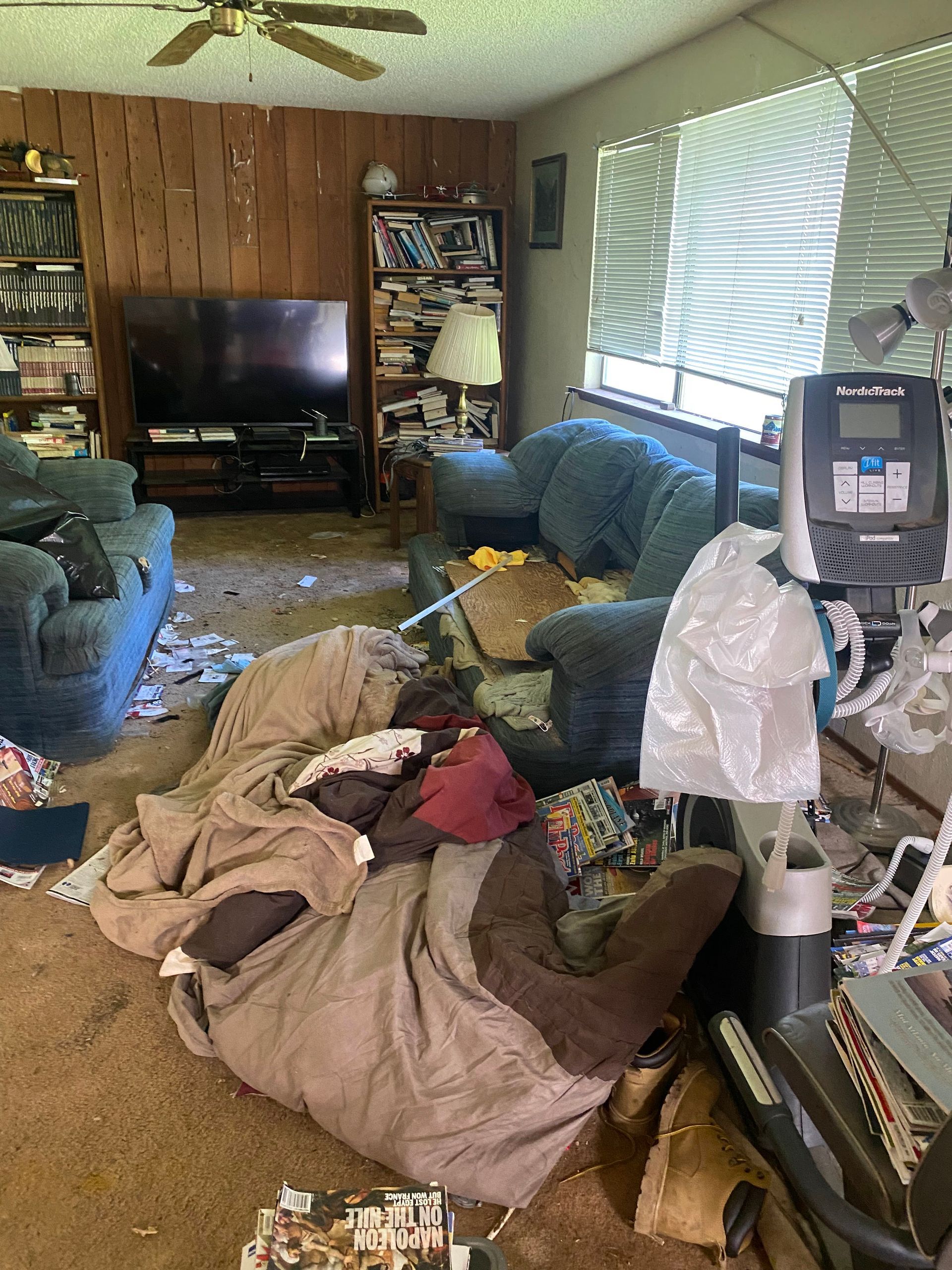 Messy living room, cluttered with trash and furniture; brown walls, blue sofa, window with blinds.