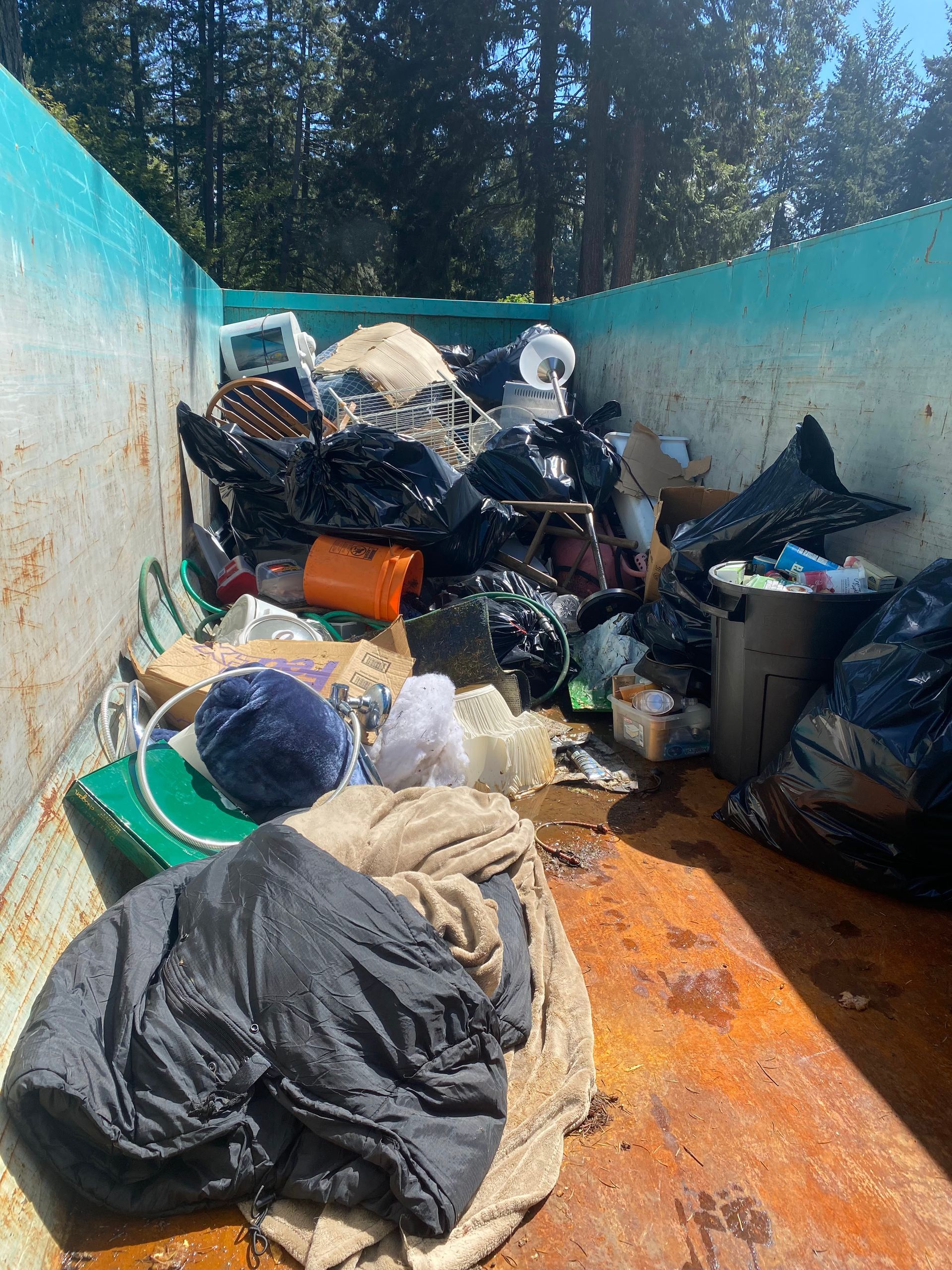 A dumpster overflowing with trash: black bags, clothes, baskets, and other debris, in a wooded area.