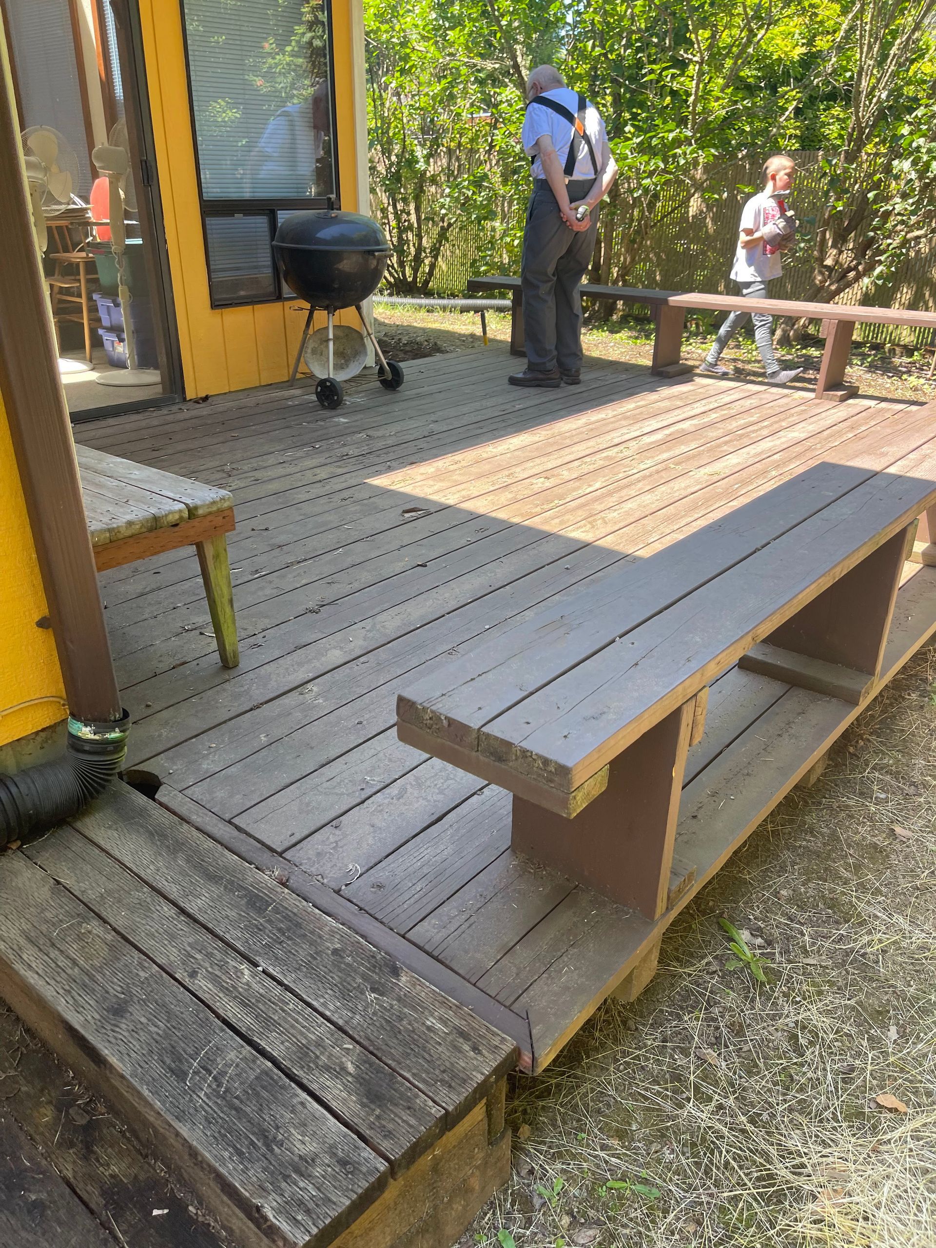 Wooden deck with built-in bench. People stand nearby. Grill and yellow building in the background.