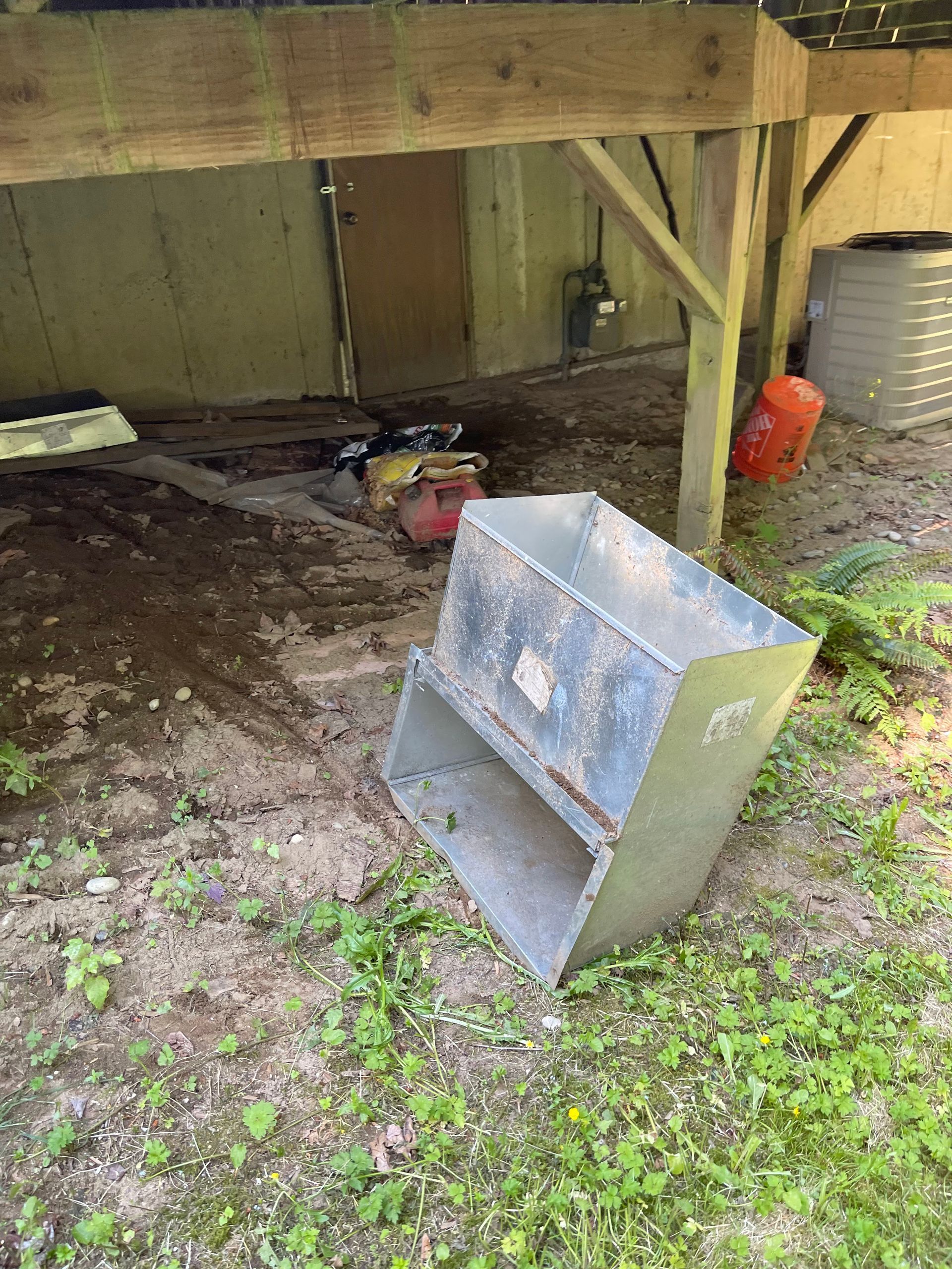 Metal bin on the ground under a wooden structure, with tools and vegetation.