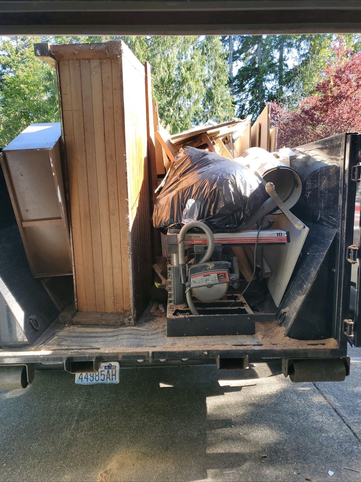 A pickup truck bed loaded with wood, tools, and debris; a radial saw is visible in the front.