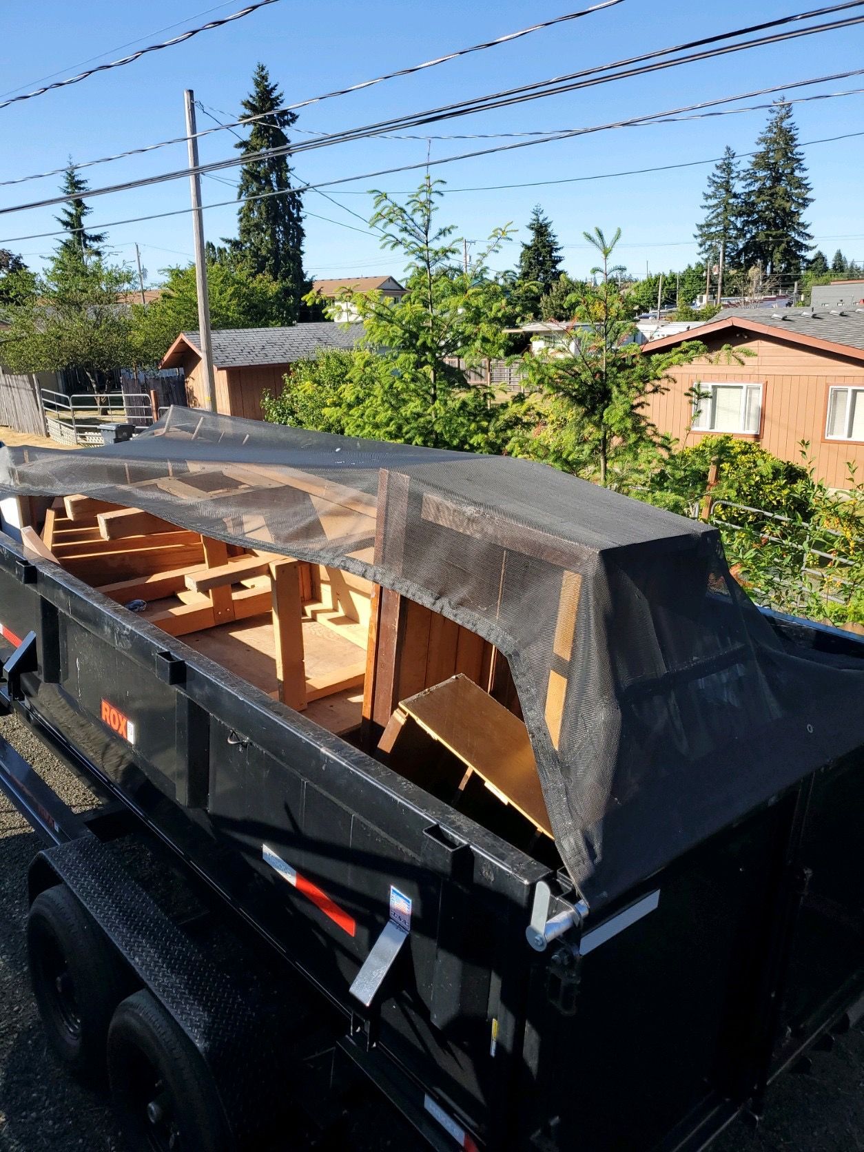 Black dumpster with a wooden frame, covered in netting, sits on a trailer, outdoors.