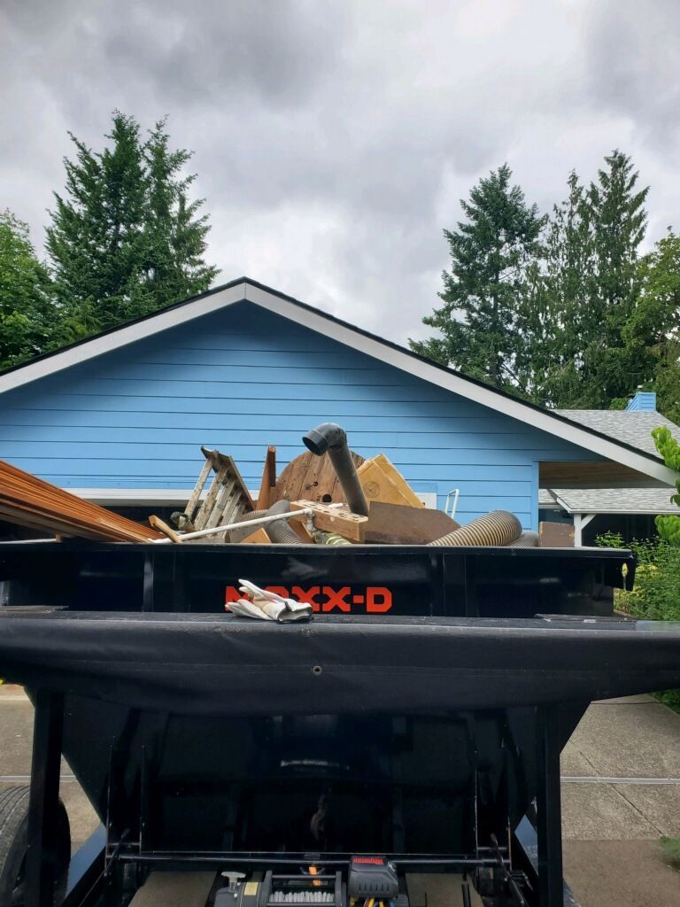 Black dumpster overflowing with debris, in front of a light blue house under a cloudy sky.