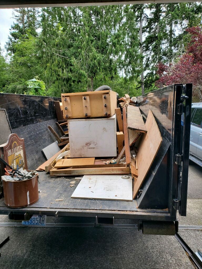 Dumpster filled with wood and debris in a driveway, trees in the background.