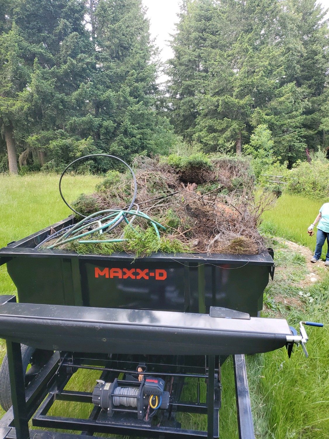 A black MAXXD trailer filled with yard waste; a bicycle wheel is visible. A person stands nearby in a grassy field.