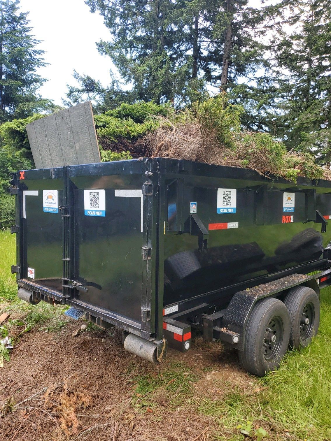 Black dumpster trailer filled with yard waste, parked on grass.