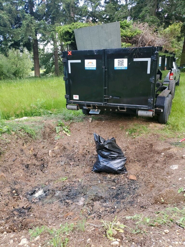 Black trash bag near dark dumpster and truck in grassy area.