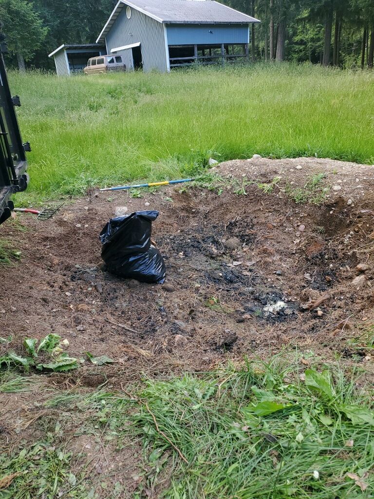 A dug-out pit with a black trash bag, dirt, and grass in front of a blue barn.