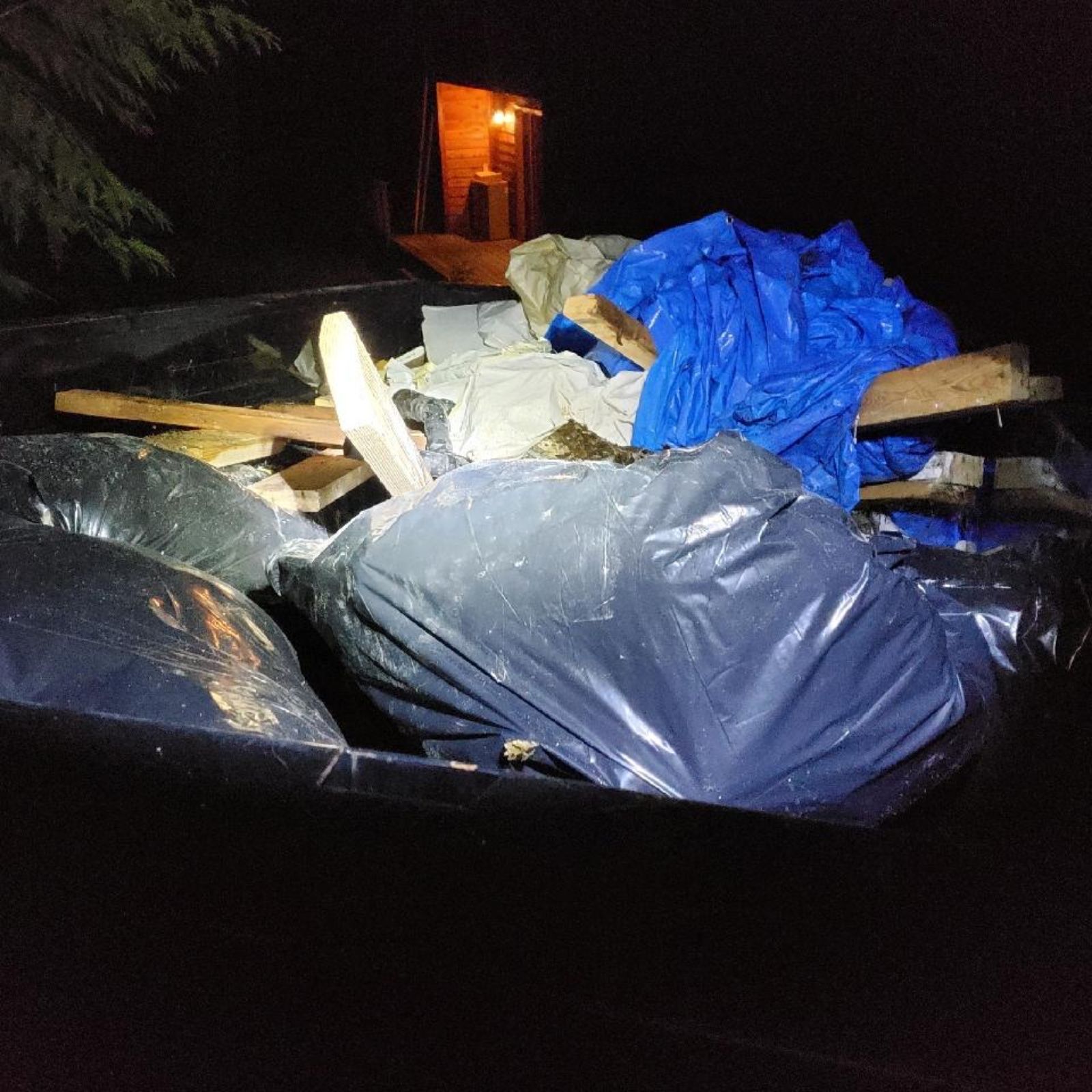 A dumpster filled with trash bags, wood, and a blue tarp at night; a structure is in the background.