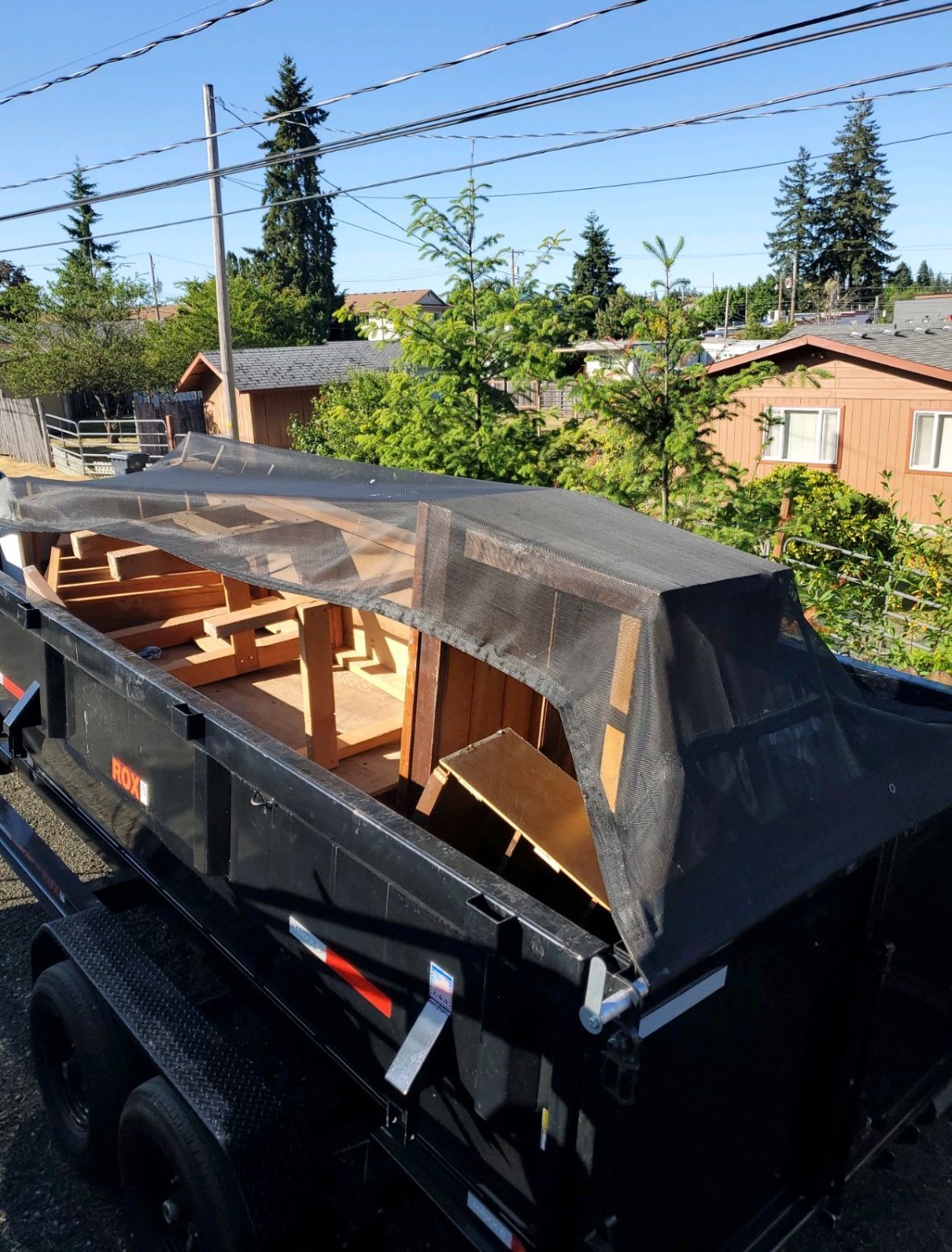 Black dumpster trailer with a wooden frame and netting on top, in a residential area.