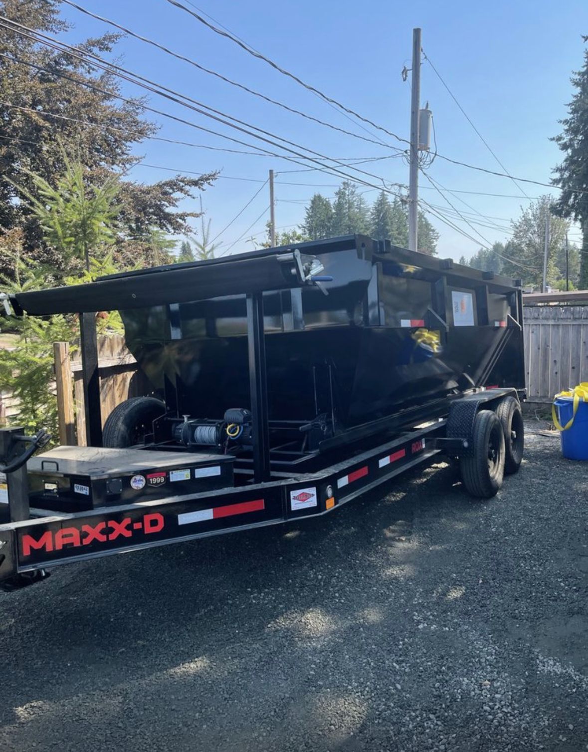 Black Maxx-D dump trailer parked on asphalt. Black container, red accents, sunny outdoor setting.