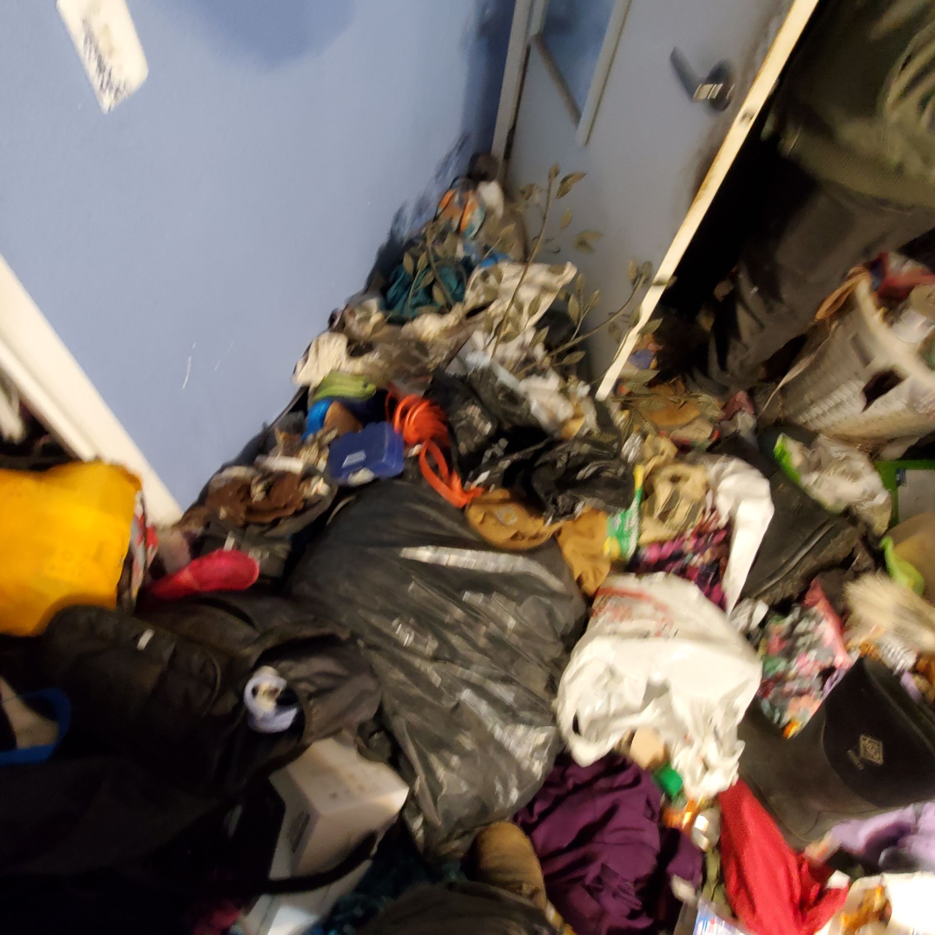 Overstuffed room with piles of clothes, trash bags, and miscellaneous items near a blue wall and doorway.